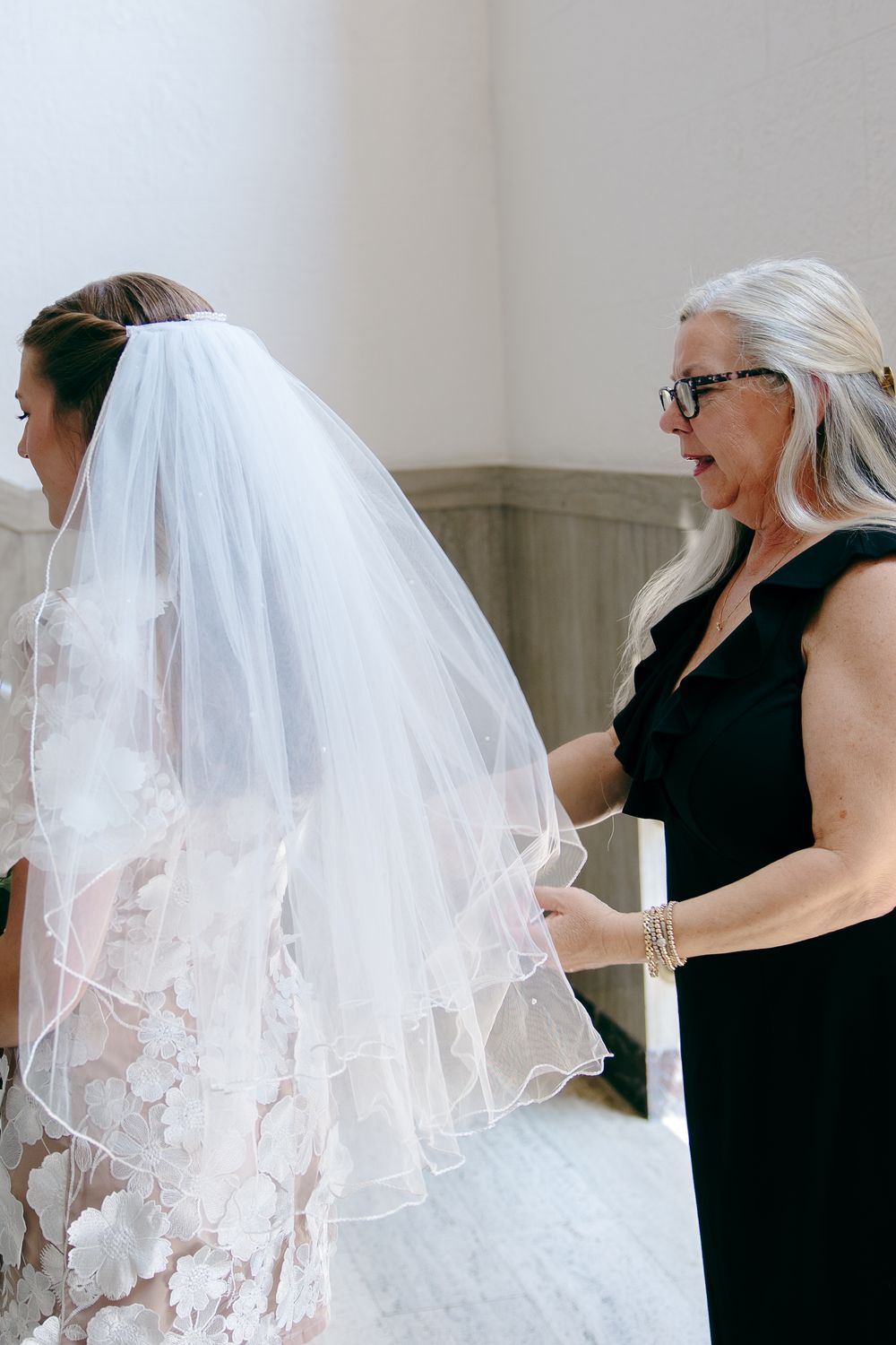 A woman in a black dress helps adjust a bride's flowing lace veil during wedding preparations.