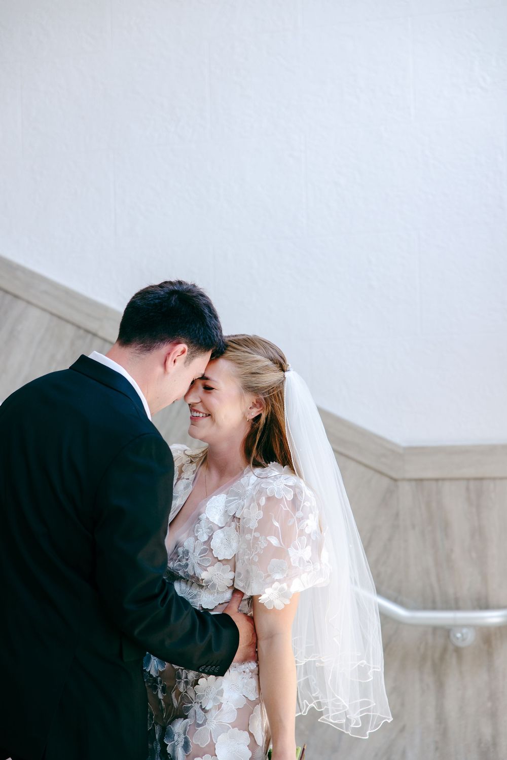 Two newlyweds share an intimate moment at their wedding, wearing black suit and floral lace dress with veil.