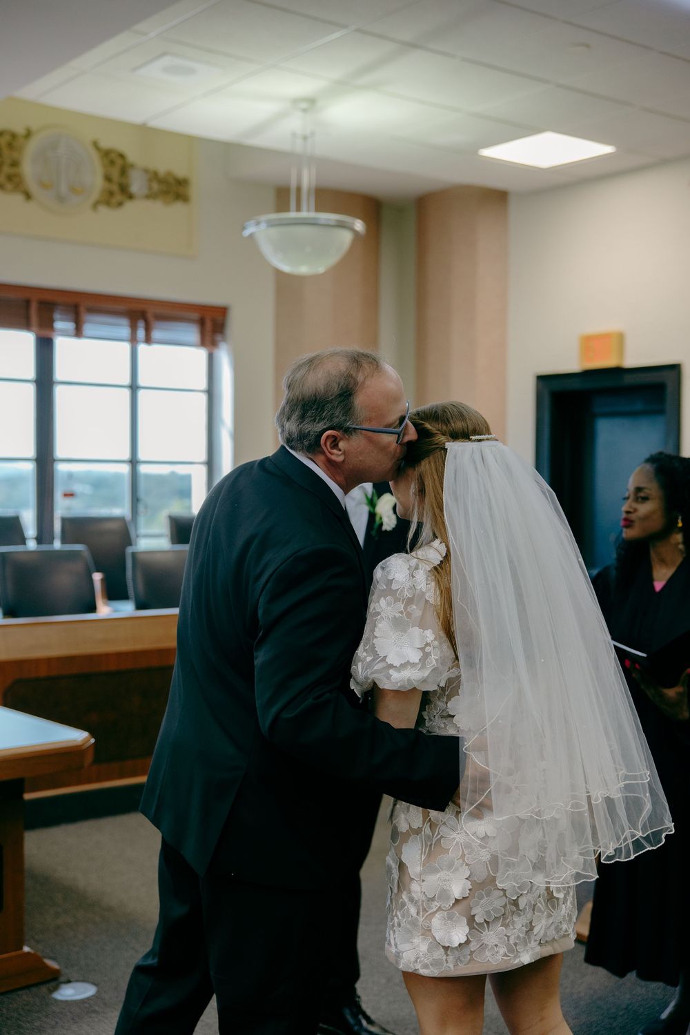 A tender moment during an indoor wedding ceremony as a bride in a short lace dress and veil receives a kiss.