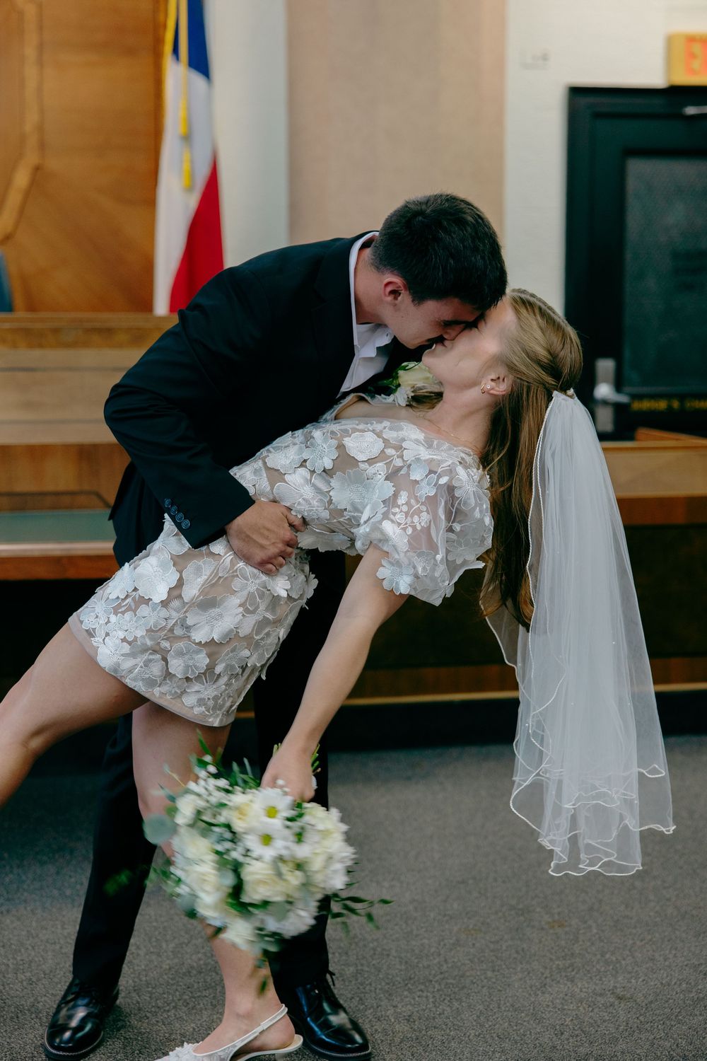 A romantic wedding dip kiss with the bride in a lace dress and veil holding a white bouquet.