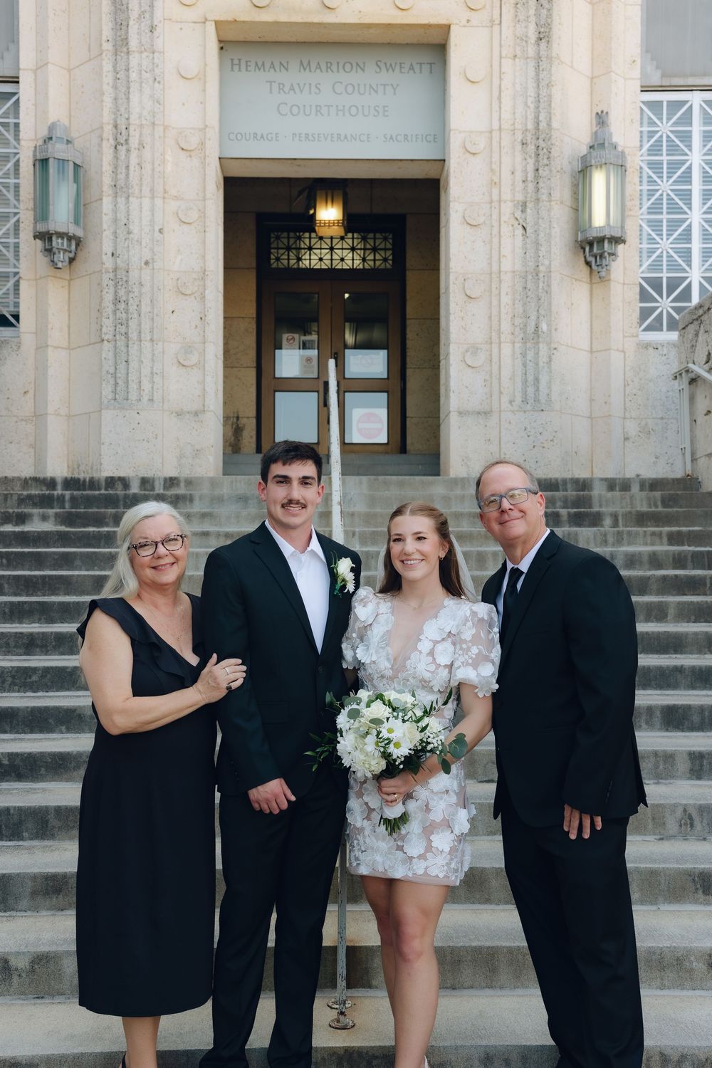 A wedding party poses on stone steps outside a historic courthouse with ornate lanterns and architecture.