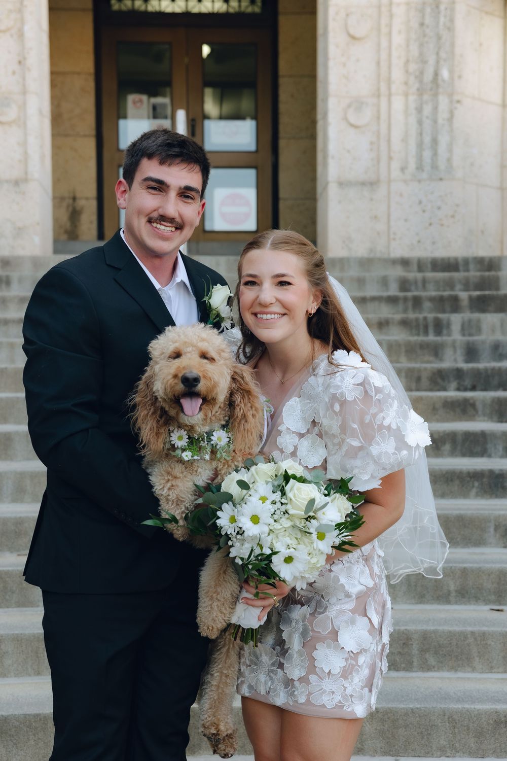 A couple in wedding attire poses on stone steps with their goldendoodle dog wearing a floral collar and white bouquet.