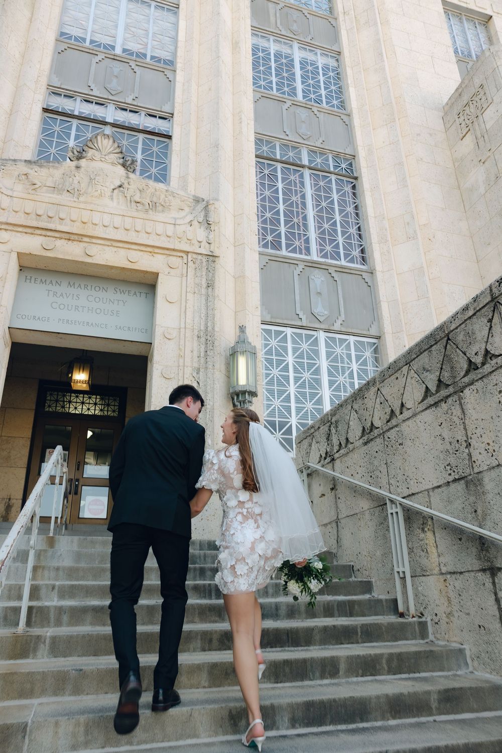 Bride and groom walking up grand stone steps of a classical building with decorative windows.