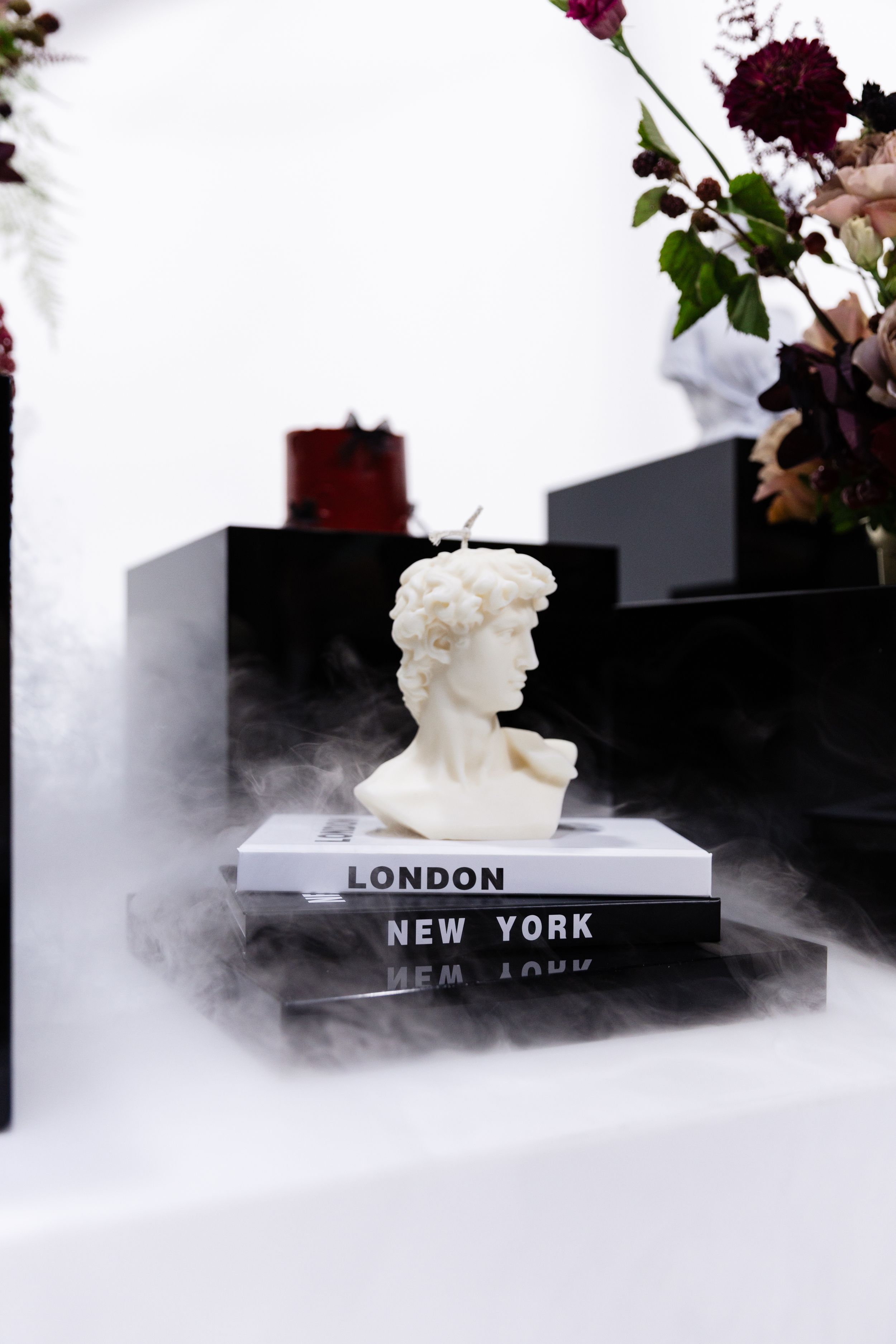 Classical bust sculpture on stacked books with misty atmosphere and floral decorations.