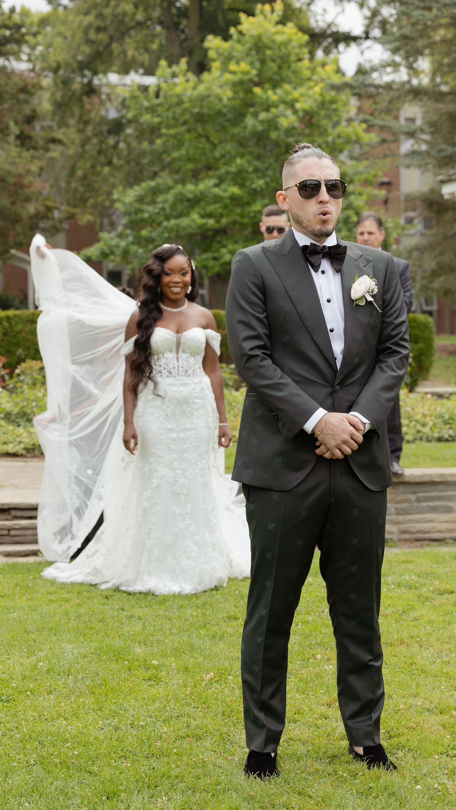 A newlywed couple in a garden, with a flowing white wedding gown and groom in a classic black tuxedo with sunglasses.