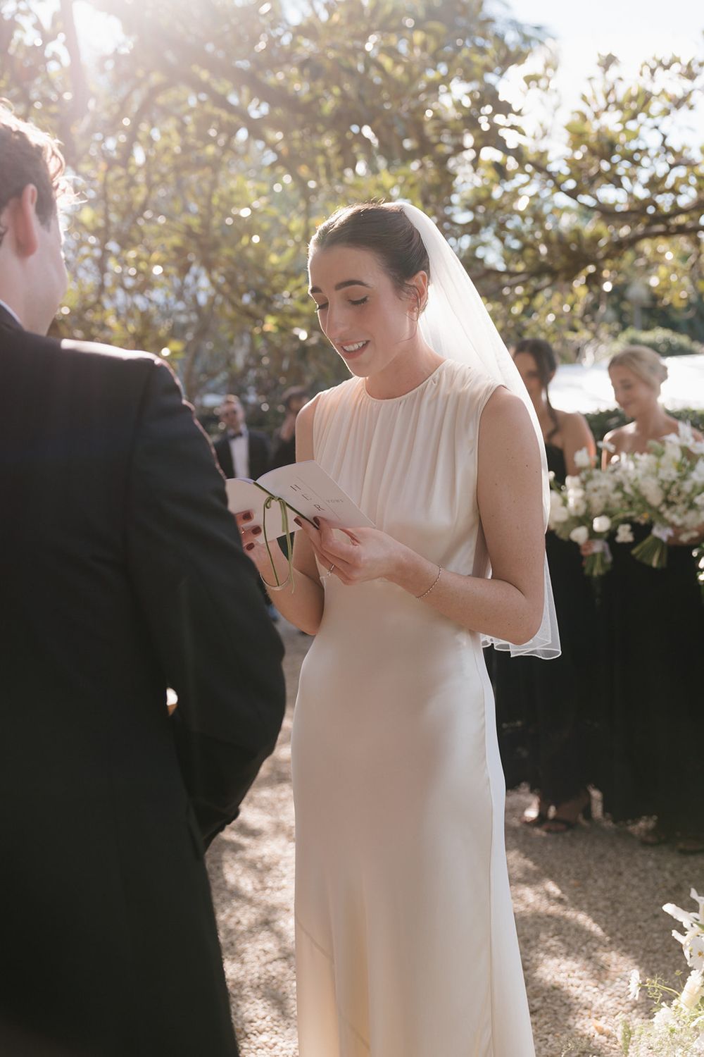 A bride in a sleek white sleeveless gown reads from a paper during an outdoor wedding ceremony in natural sunlight.