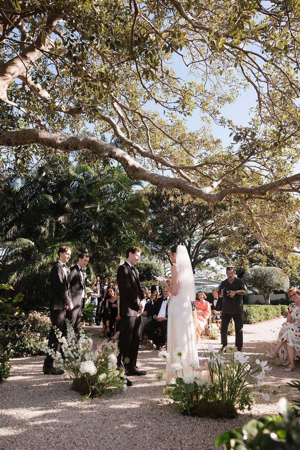 A garden wedding ceremony takes place under a large spreading tree on a sunny day with floral arrangements decorating the path.