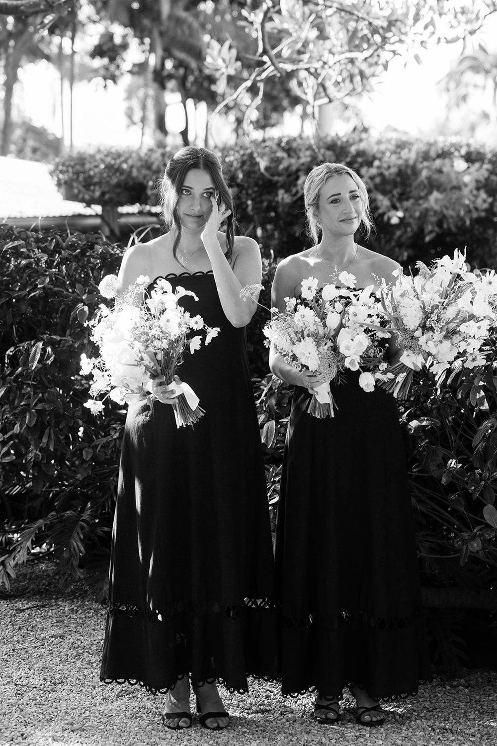Two bridesmaids wearing elegant black dresses hold beautiful white floral bouquets while standing together in a garden setting.