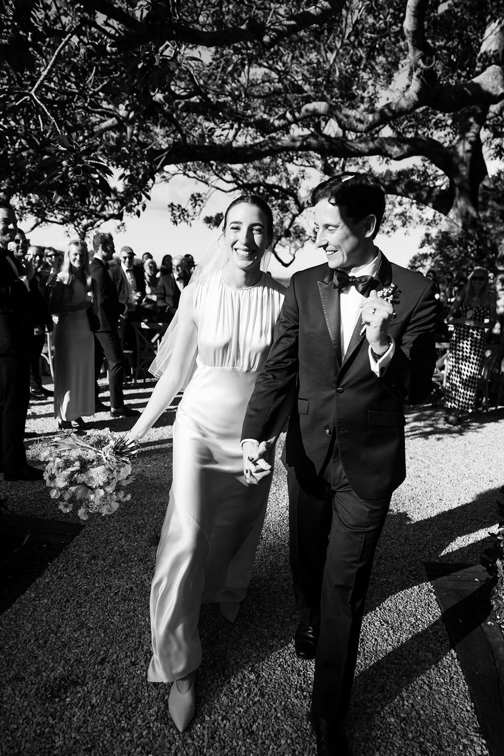 A black and white photo of a joyful wedding couple walking together outdoors under a tree at their outdoor ceremony.