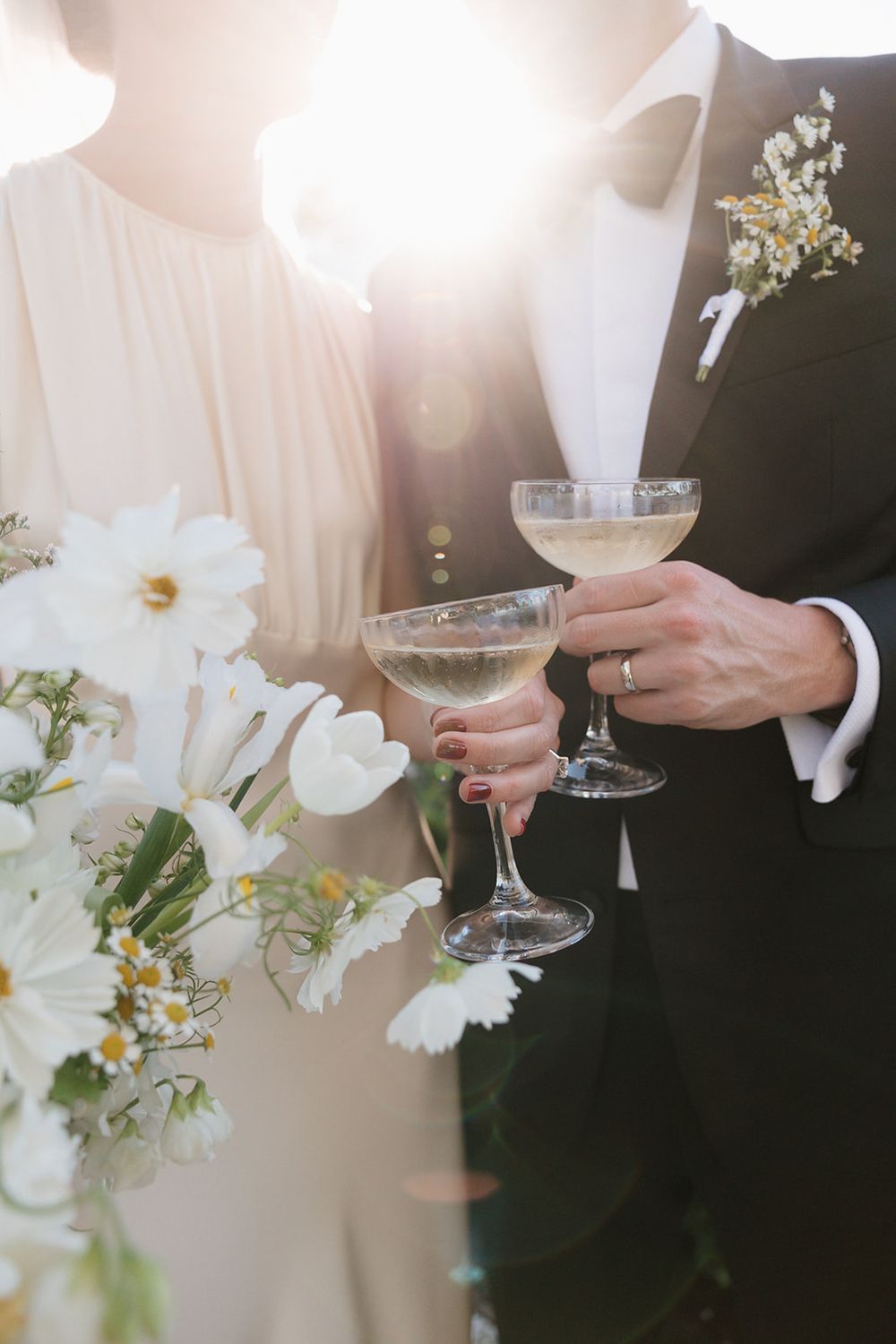 A romantic close-up of champagne glasses toasting at a wedding with white flowers and sunlight in the background.