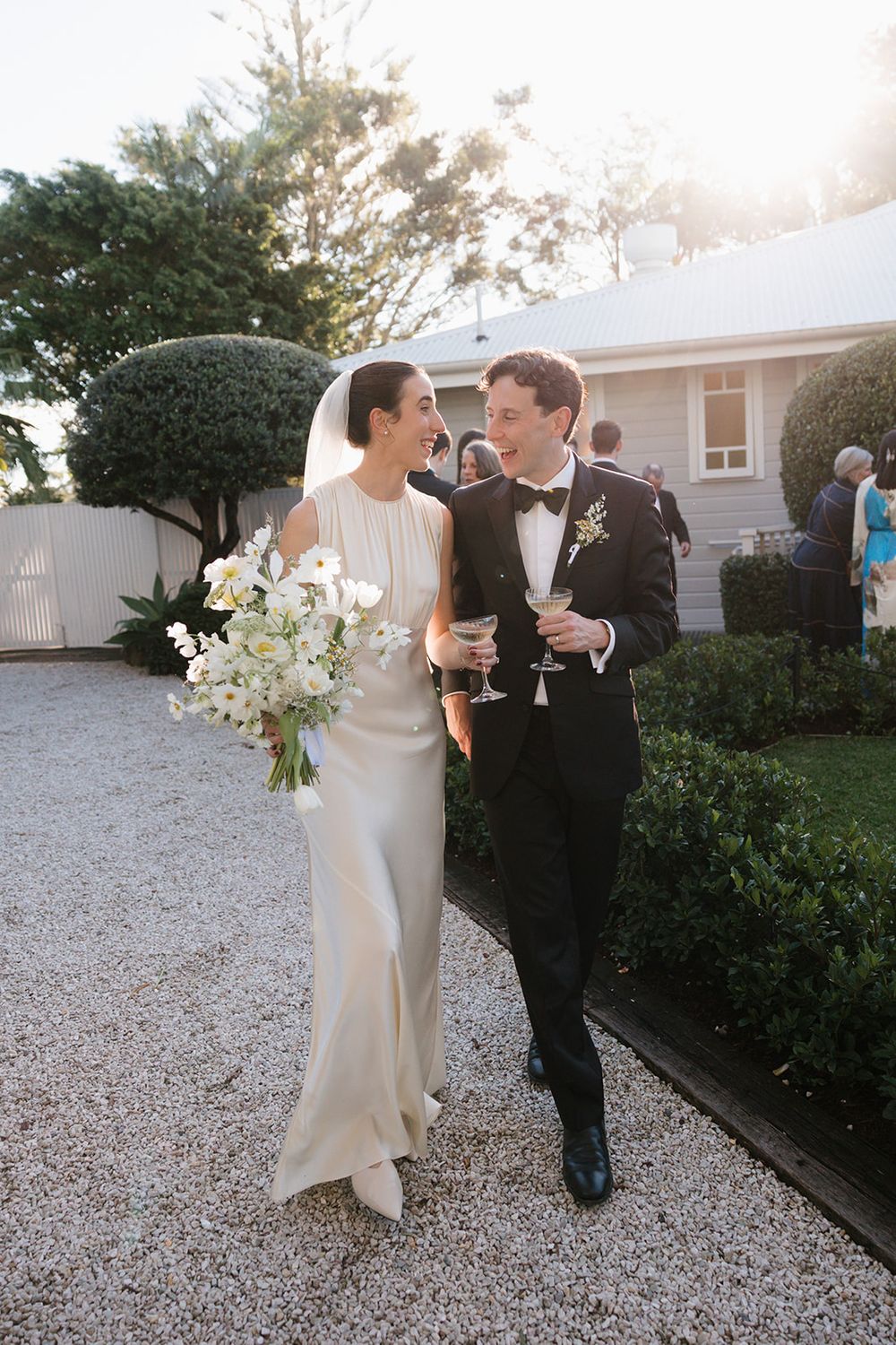 A couple walking together on a gravel path while holding champagne glasses at a sunset wedding celebration.