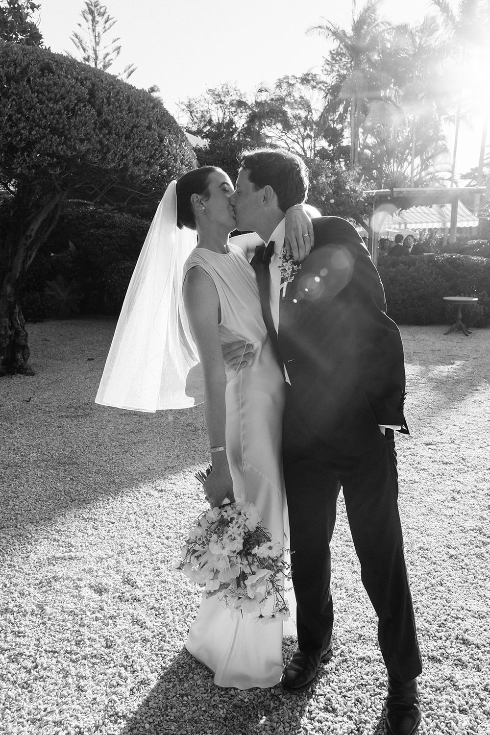 A romantic black and white wedding photo showing a couple sharing a kiss on a garden path with palm trees in the background.