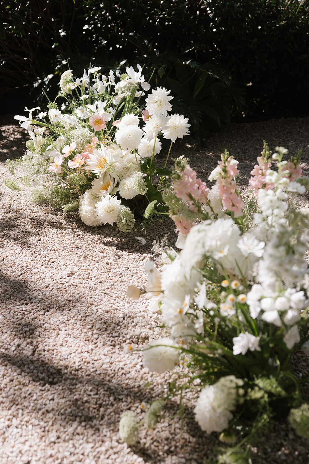 Delicate white and pink floral arrangements line a gravel pathway with soft shadows creating a romantic garden setting.