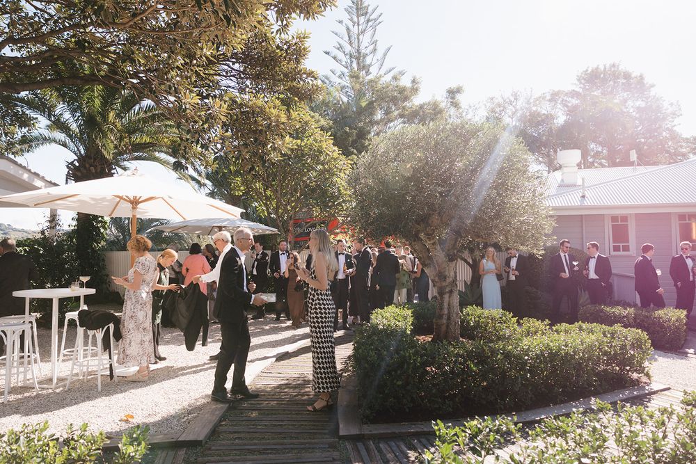 A sunlit outdoor event venue with guests gathering under white umbrellas and decorative trees in a garden setting.