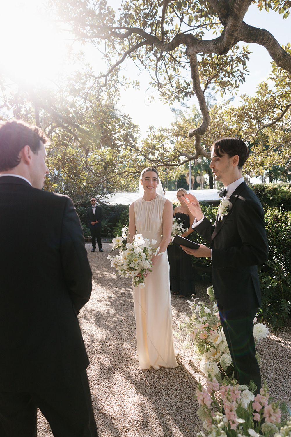 A sun-drenched outdoor wedding ceremony takes place under a tree with white floral decorations along the garden path.