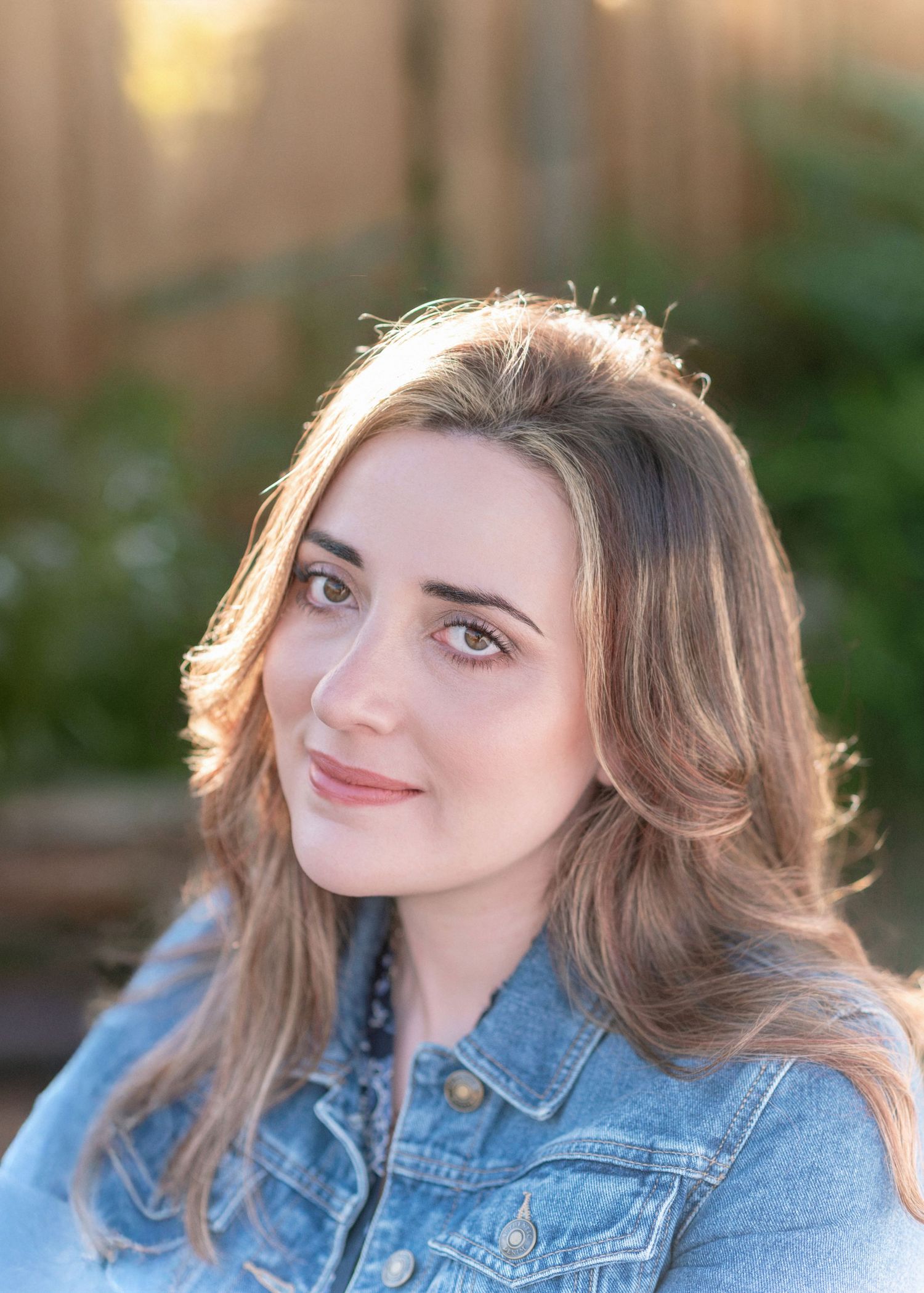 Person wearing denim jacket smiles softly in warm outdoor lighting with soft focus background.