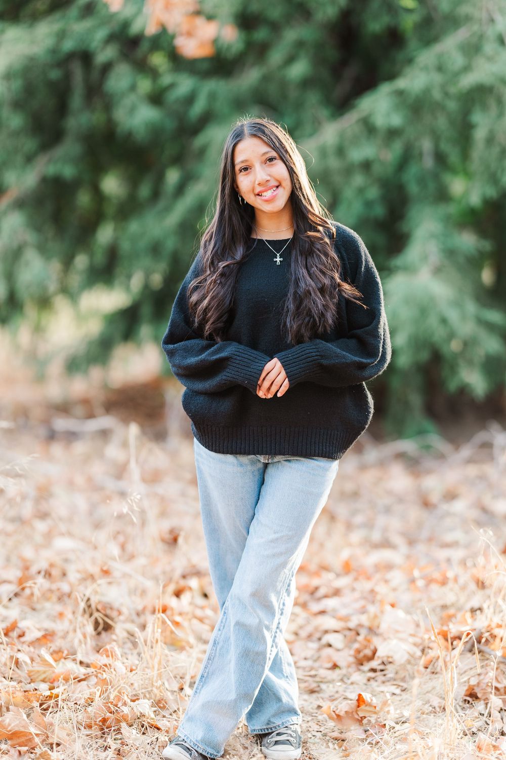 Person wearing a black sweater and light blue jeans poses outdoors in an autumn setting with fallen leaves.