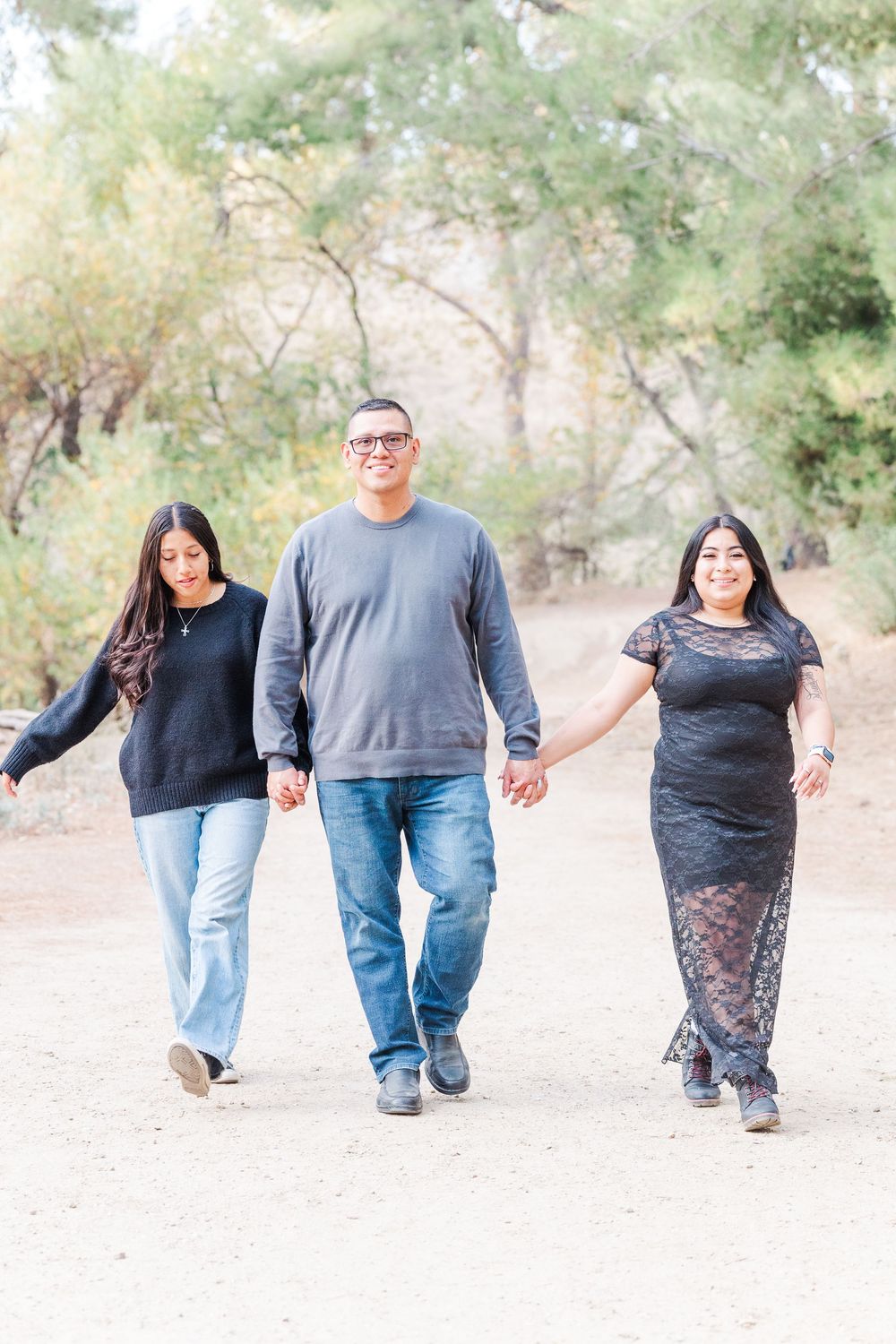 Three people holding hands while walking together on a dirt path surrounded by green trees and natural scenery.