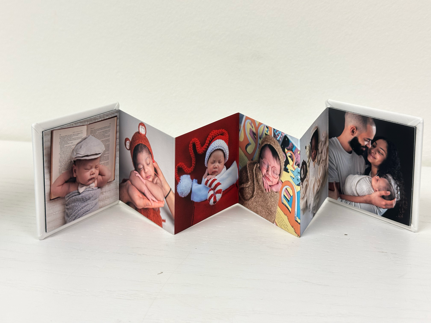 Accordion-style mini photo book displaying baby milestone photos, standing upright on a white surface.