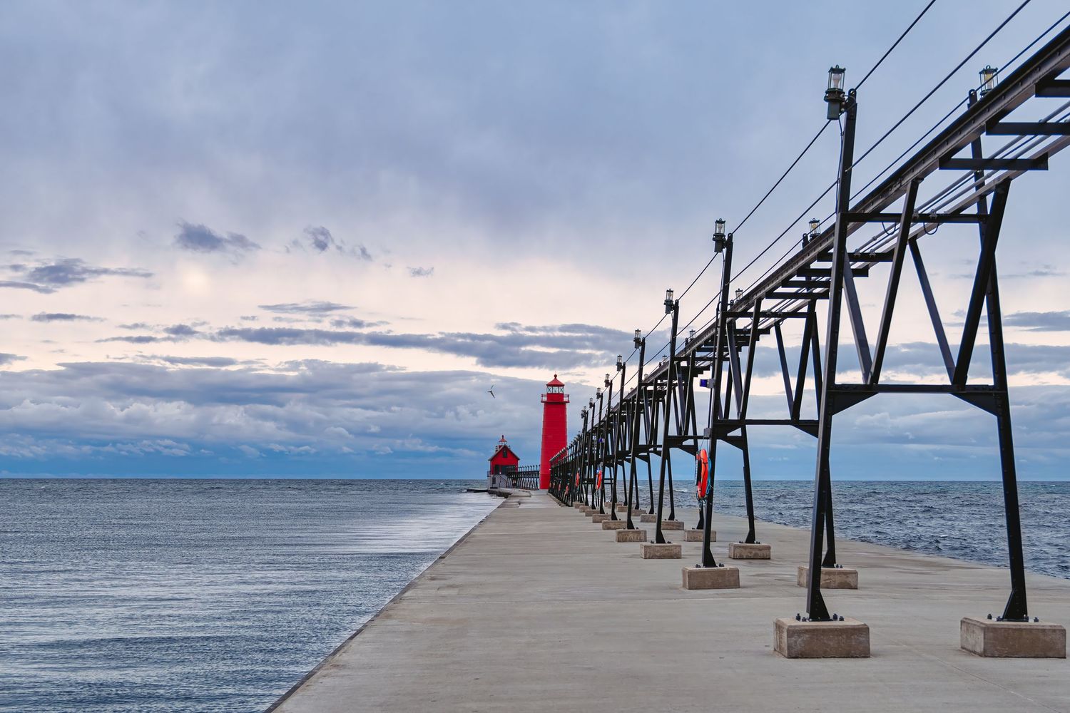 A red lighthouse stands at the end of a long pier under dramatic Michigan skies, captured by Carolina Waldroff Photography.