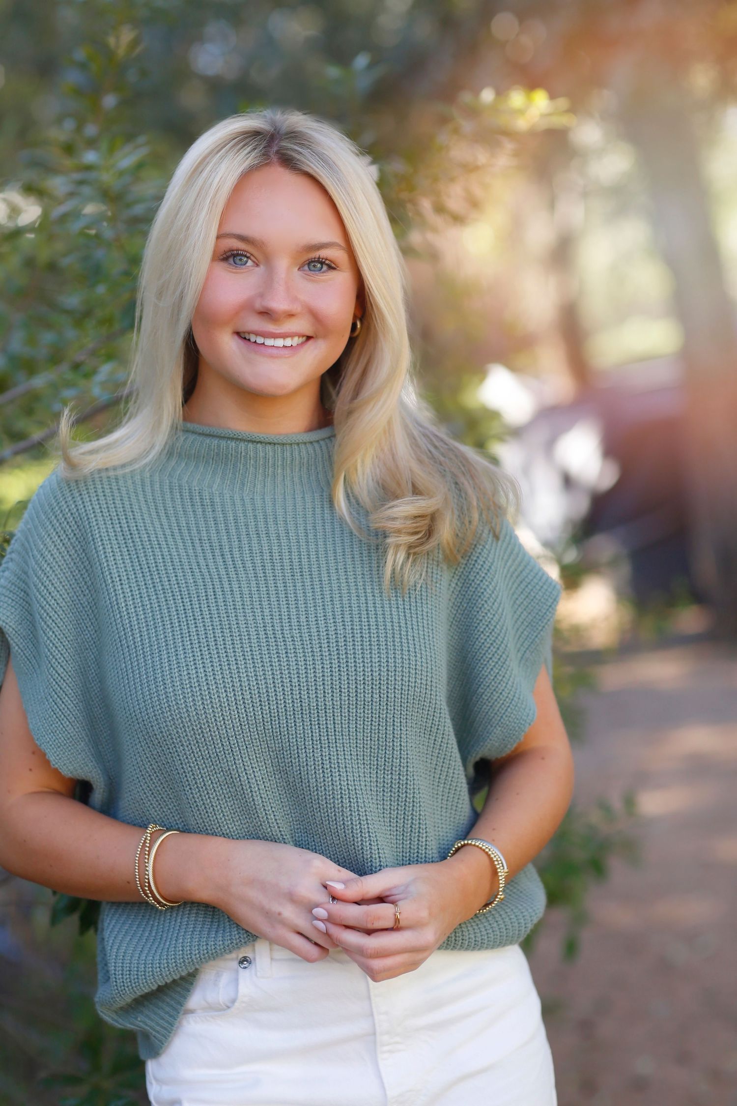 Portrait of someone wearing a sage green sweater and white pants standing outdoors with natural lighting and foliage in background.