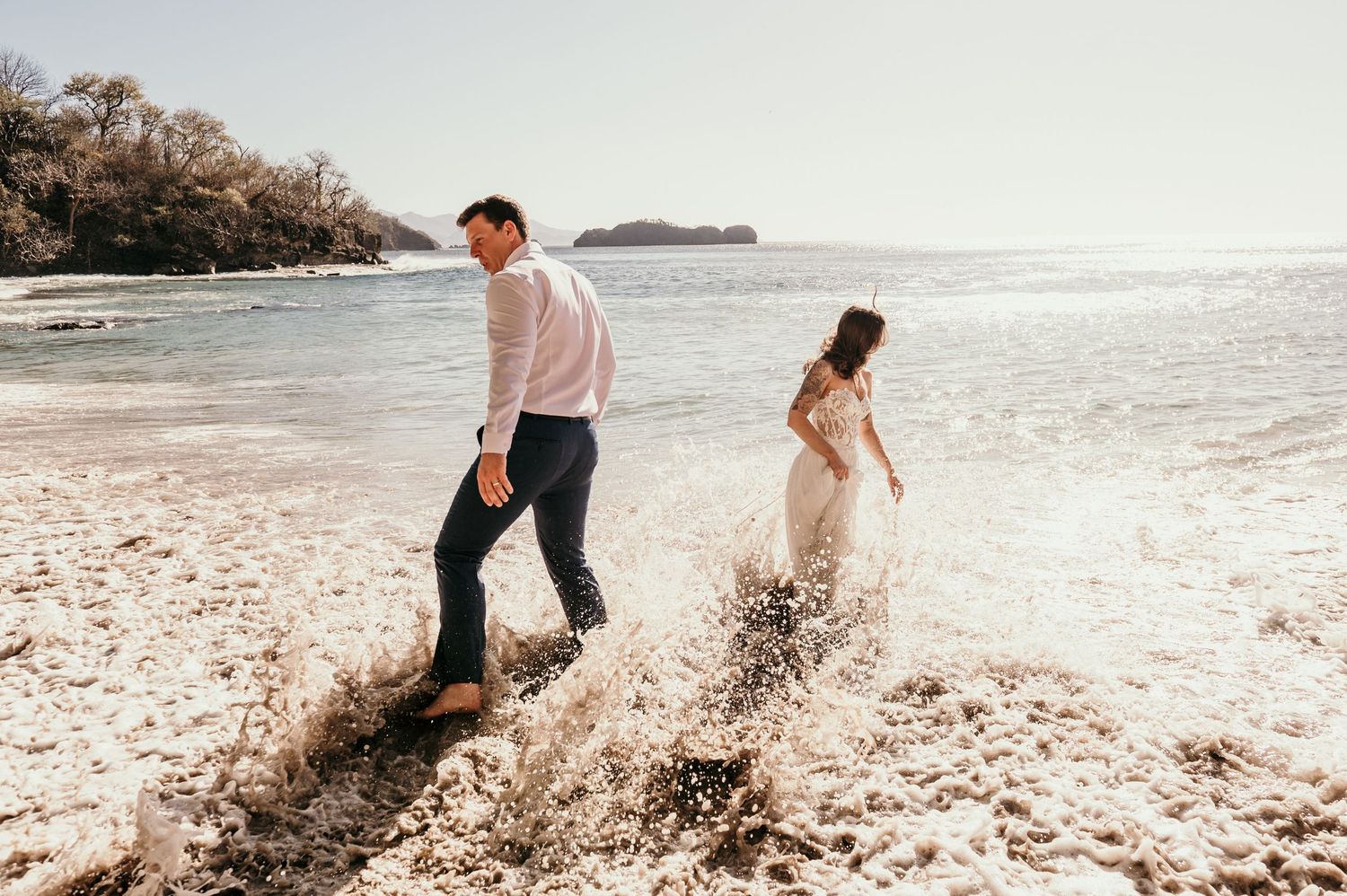 Two people splashing through waves along a sunlit beach shore with mountains visible in the background.