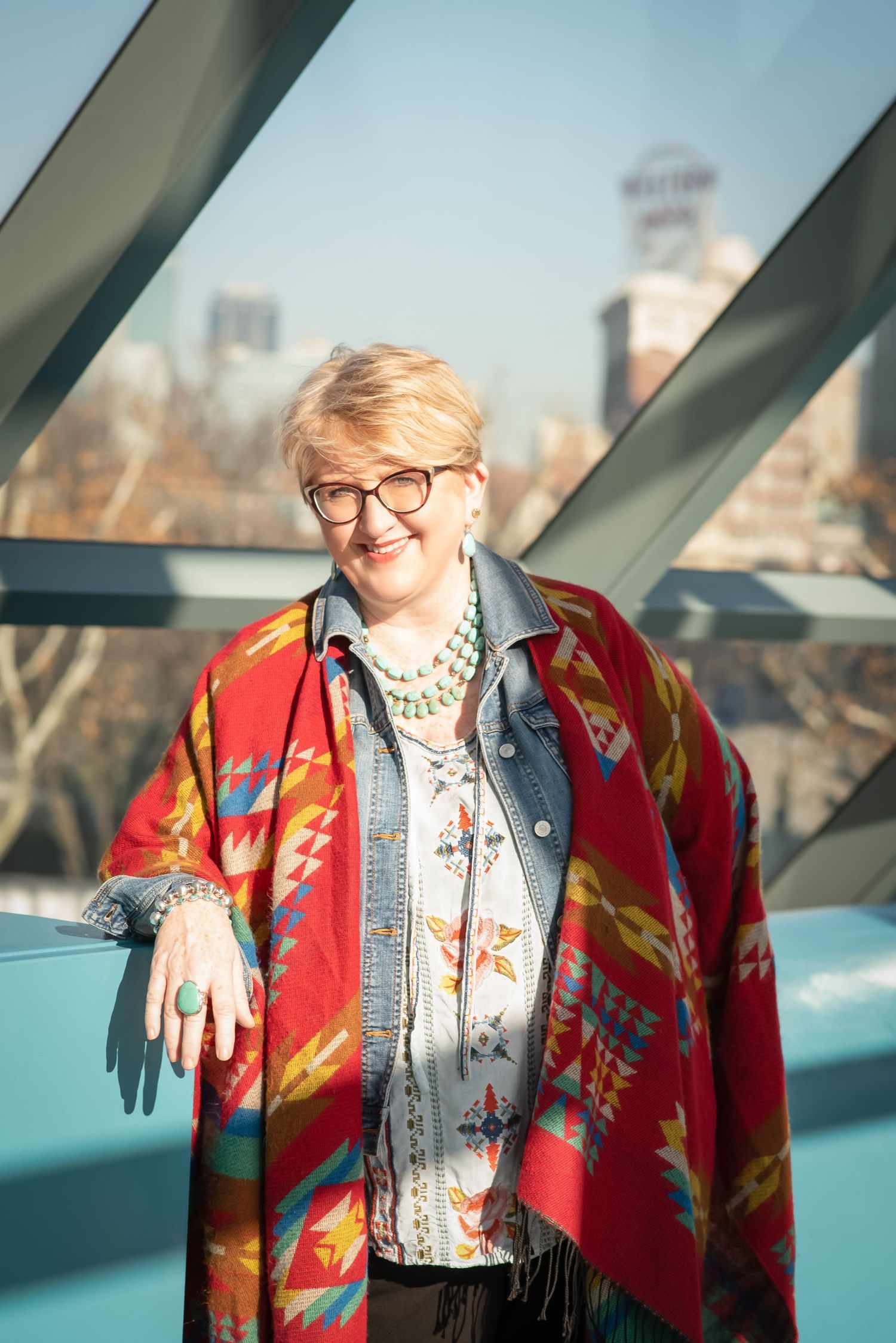 Person wearing colorful southwestern pattern shawl and glasses against city backdrop.