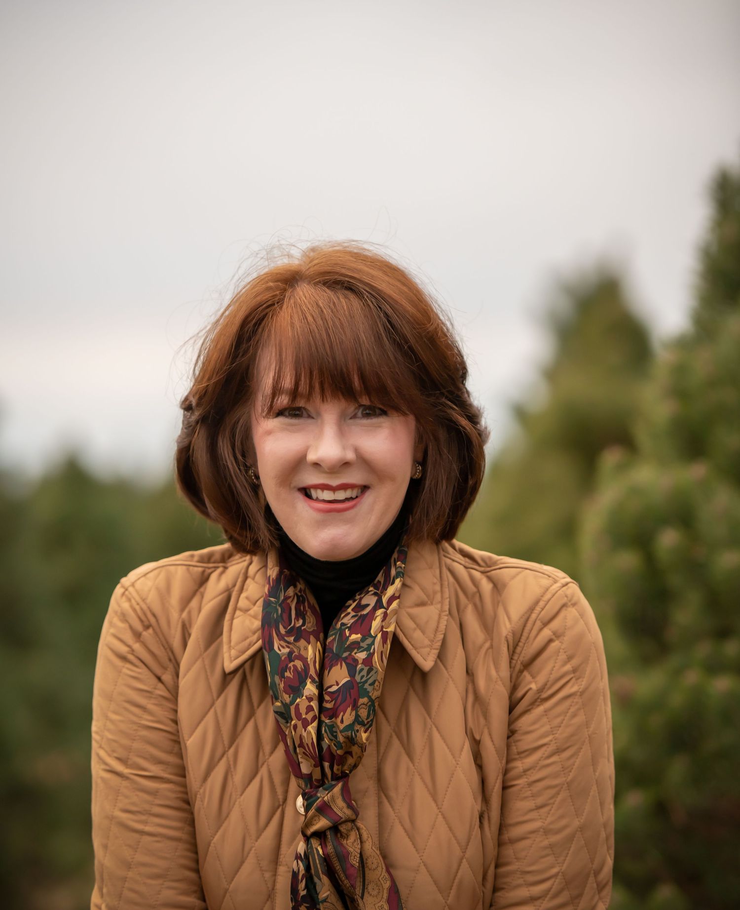 Woman in quilted tan jacket and patterned scarf smiling outdoors.