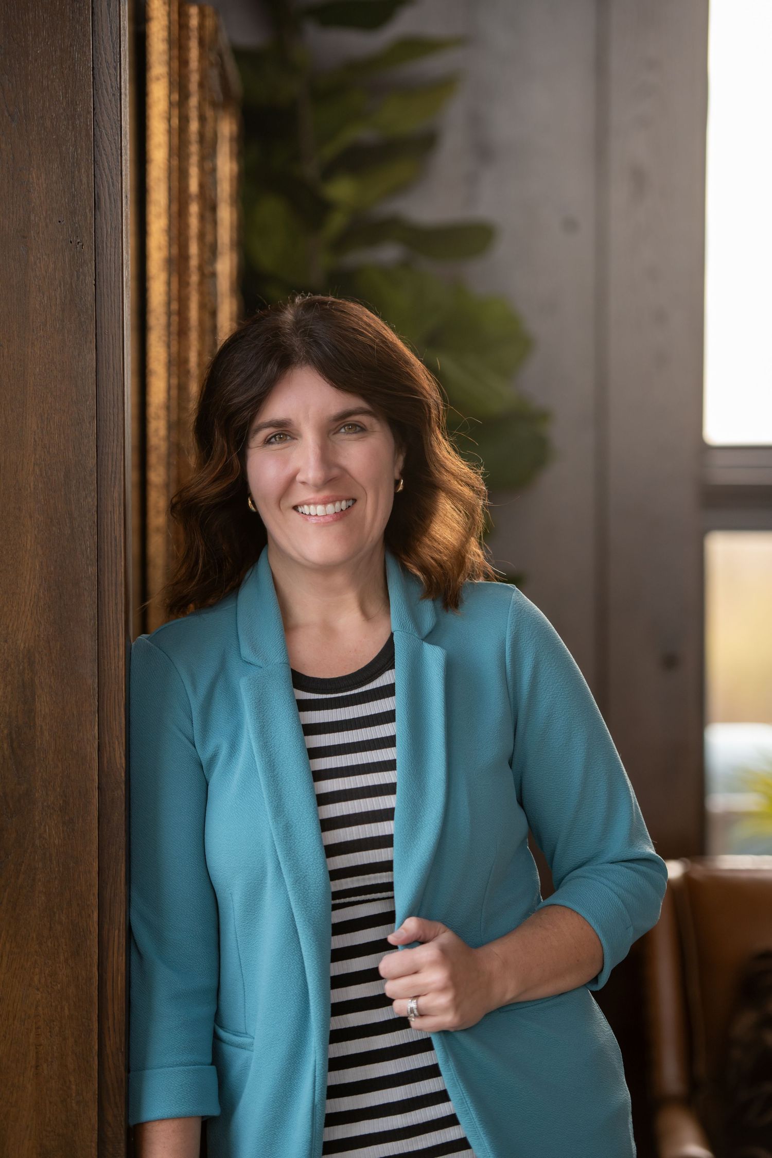 Woman in turquoise blazer and striped shirt smiling warmly in office setting.