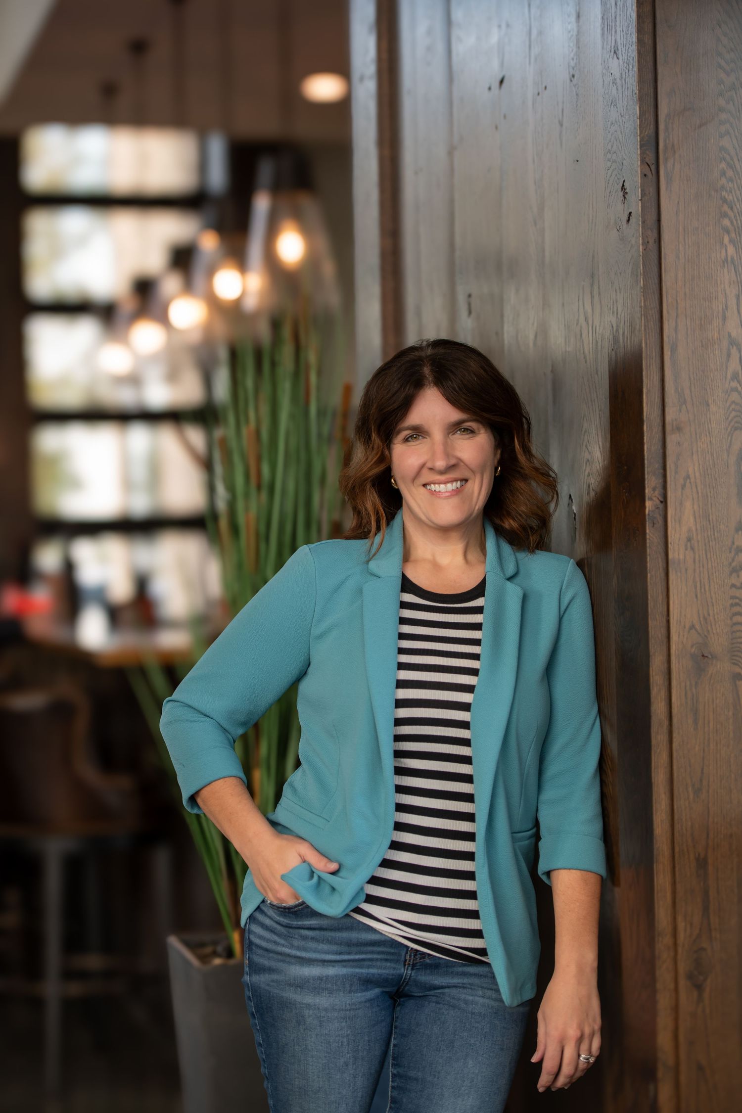 Woman in casual business attire posing against wooden wall with hanging lights.