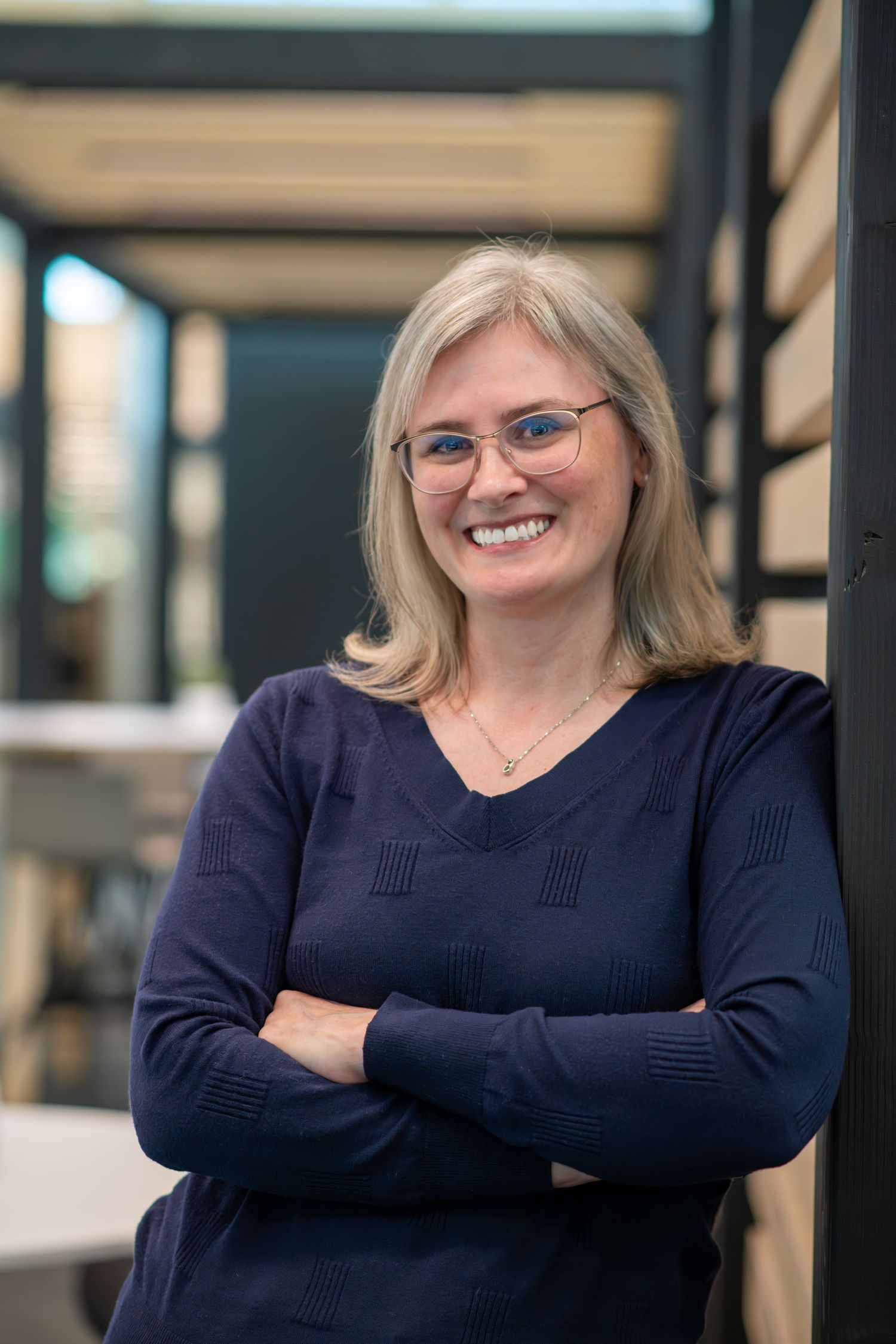 Professional woman in navy sweater and glasses standing with arms crossed in office setting.