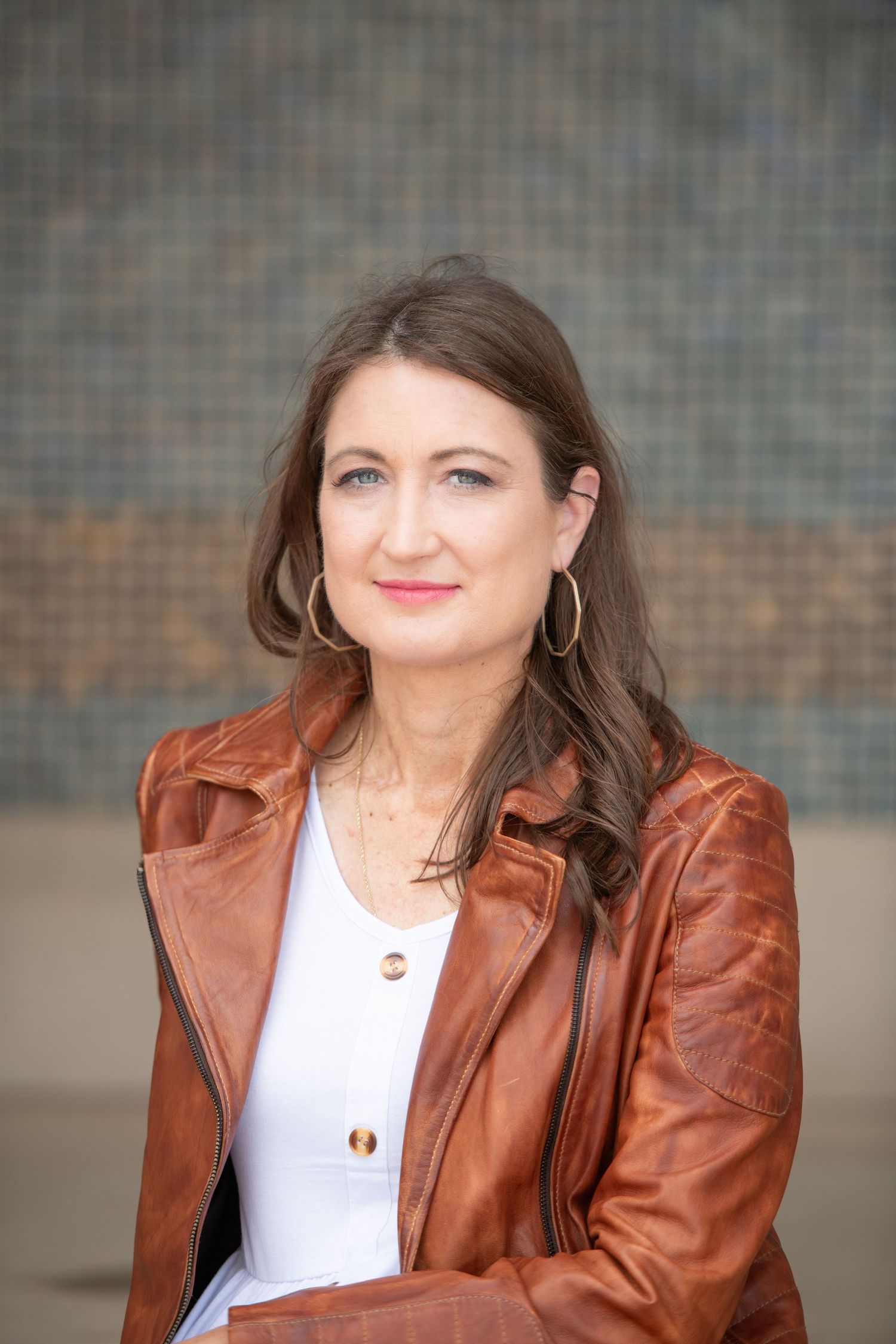 Portrait of woman wearing brown leather jacket and white blouse against industrial background.