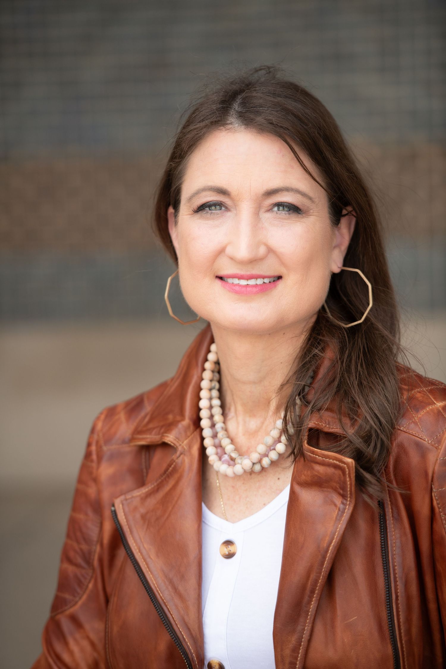 Professional headshot of woman in leather jacket with pearl necklace and hoop earrings.