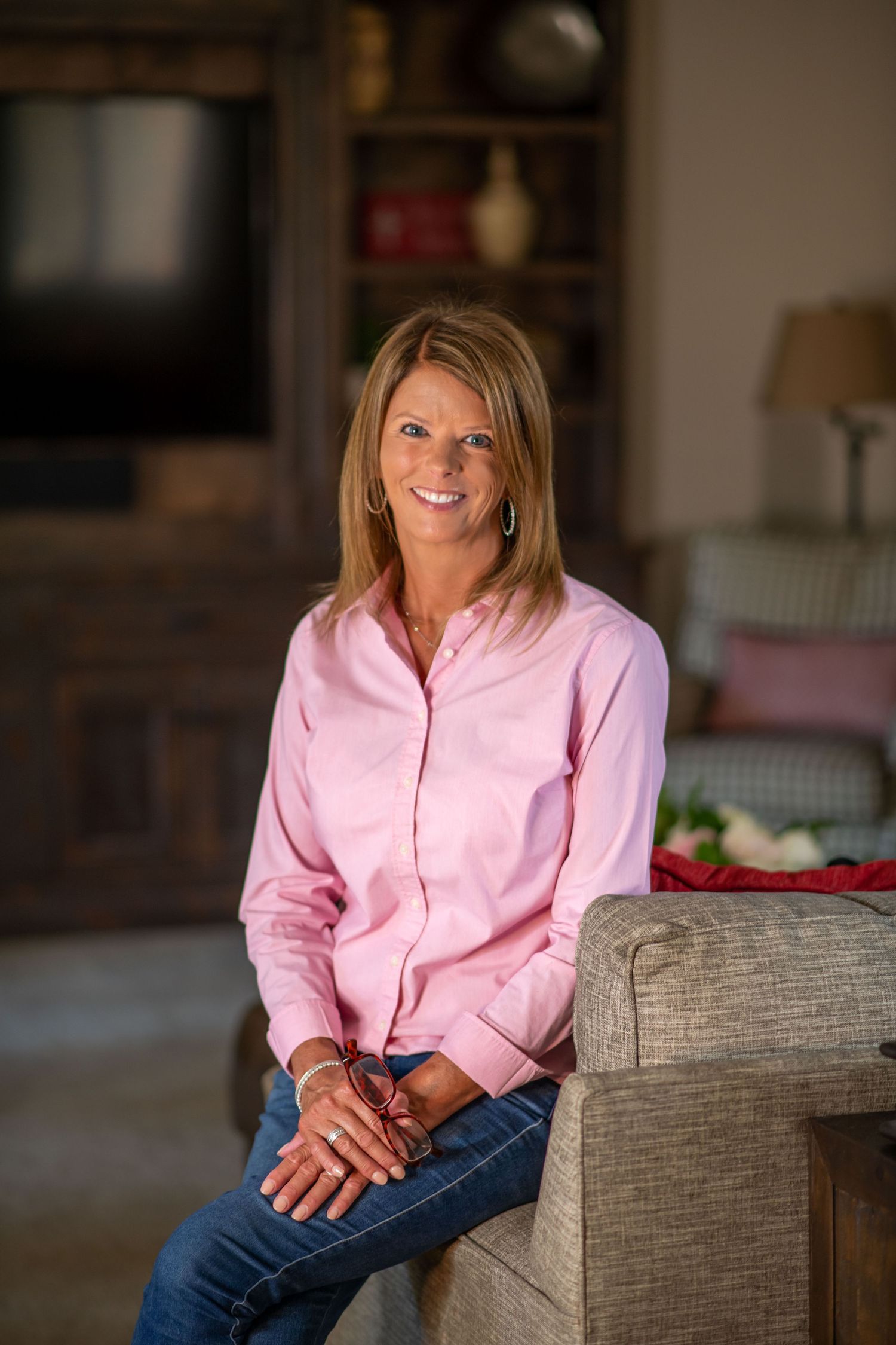 Woman in pink button-down shirt and jeans sitting casually on couch in living room.