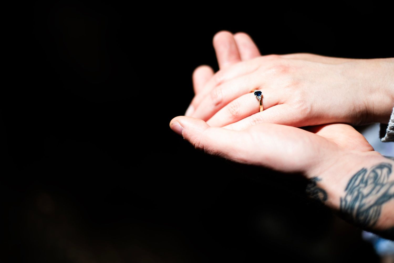 Tattooed hands gently hold a gold and silver engagement ring against a dark background.