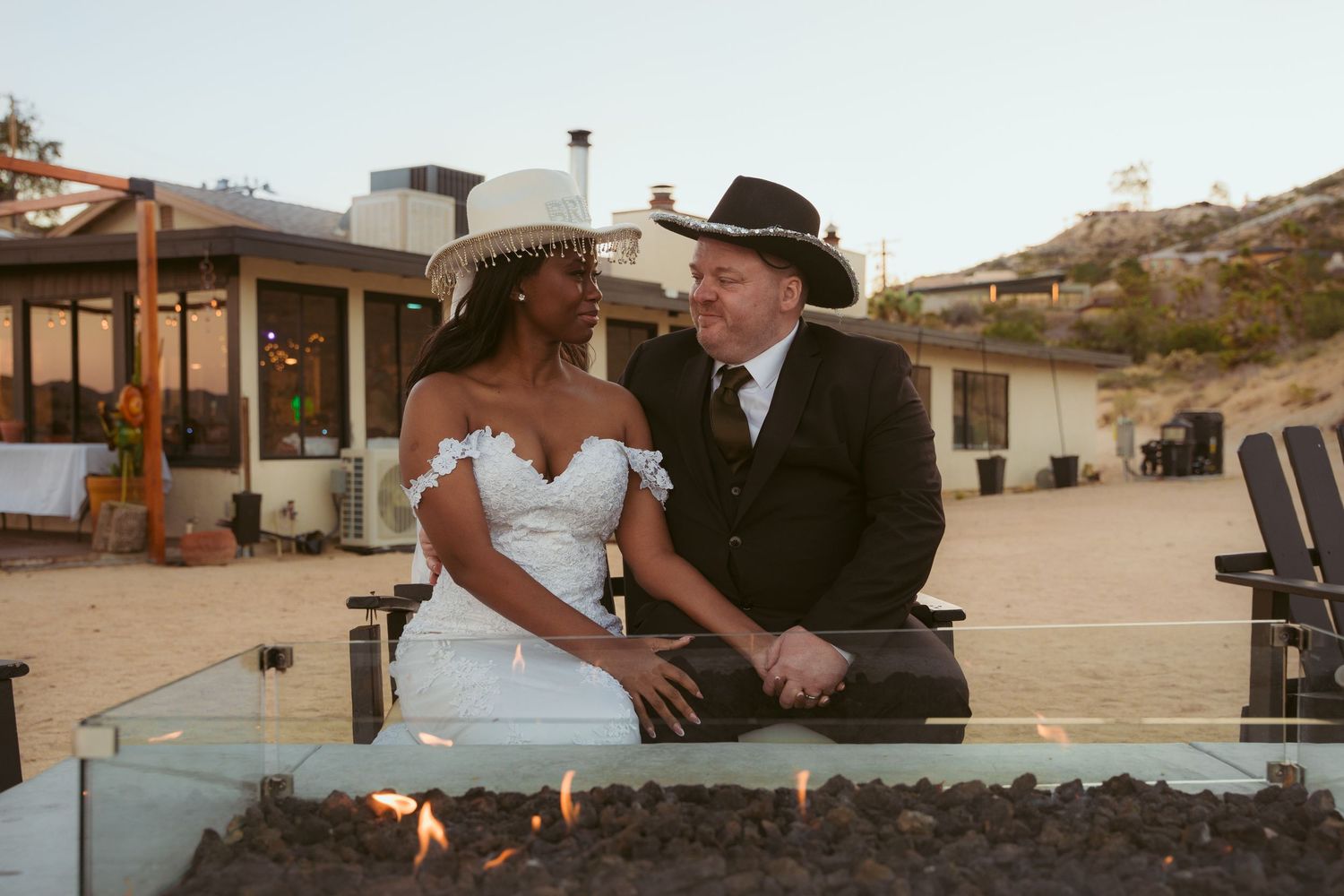 A couple sits together by an outdoor fire pit at a desert venue during a wedding celebration.