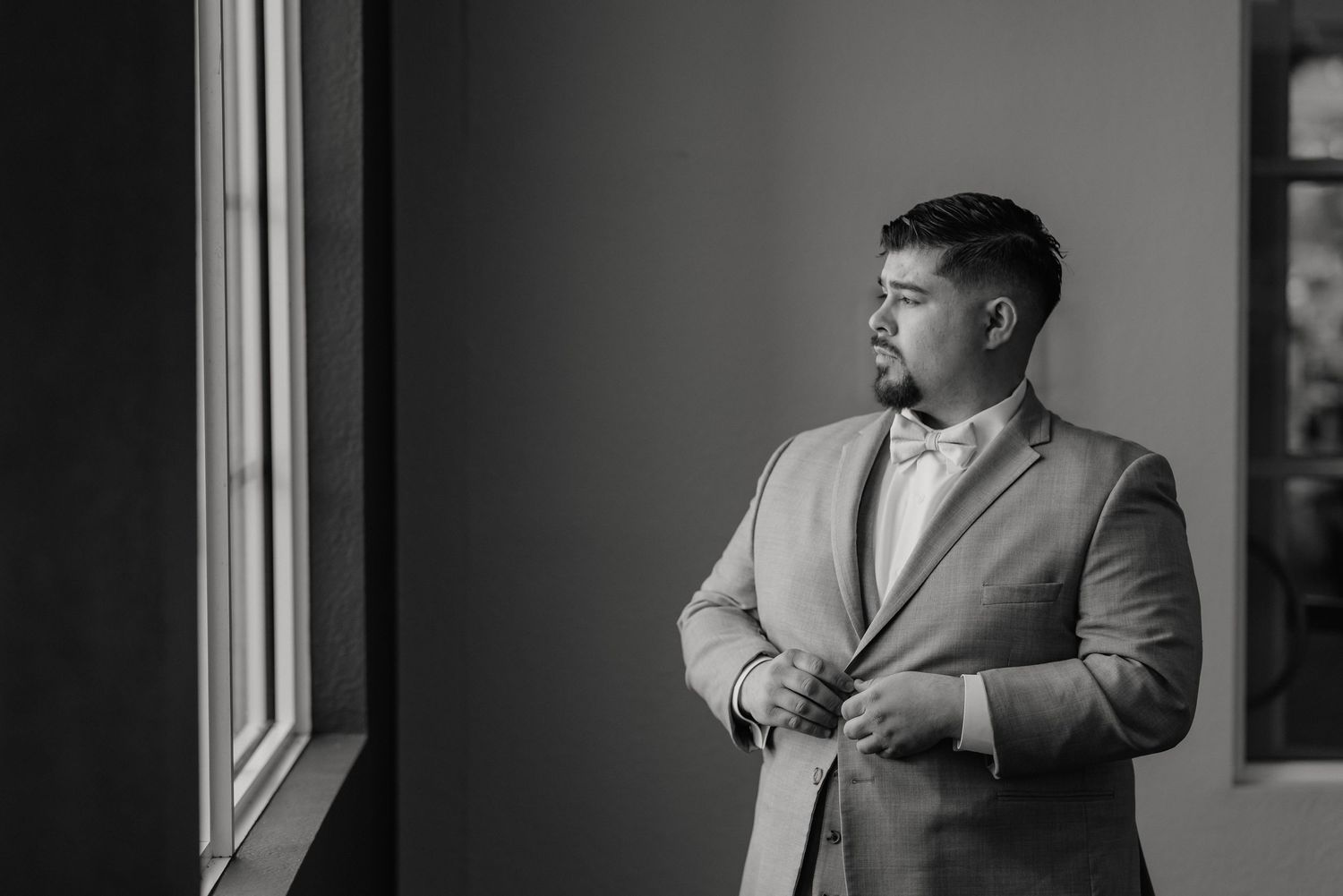A groom in a light gray suit adjusts his bowtie while gazing thoughtfully out a window in black and white.