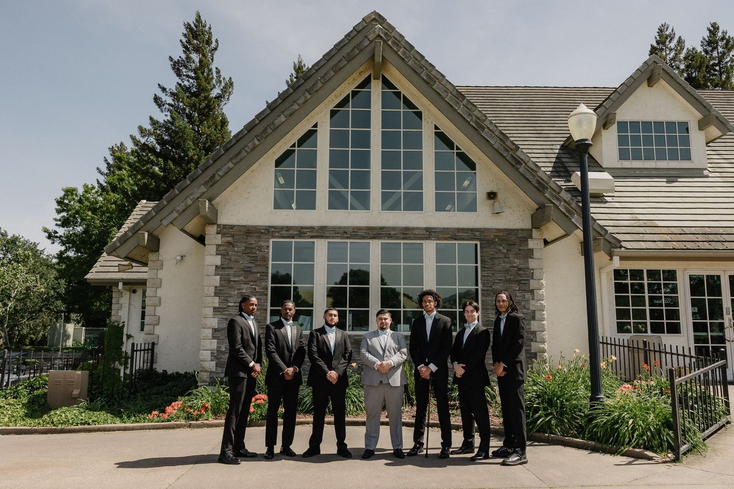 Wedding party stands outside an elegant stone and stucco house with large peaked windows and green landscaping.