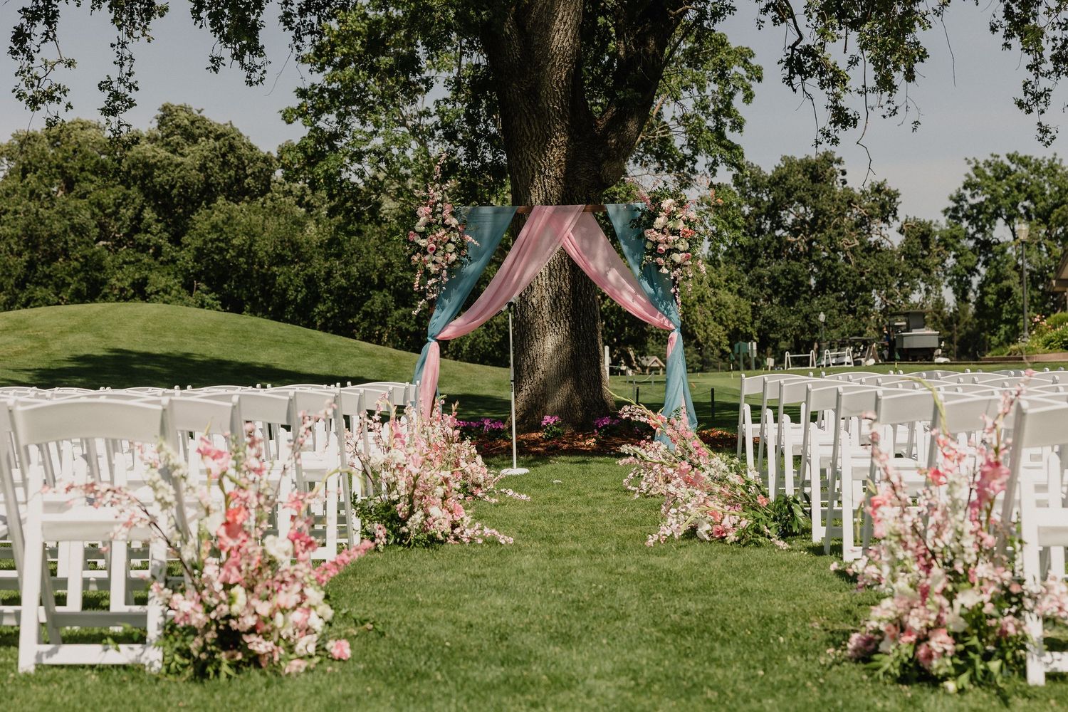 Outdoor wedding ceremony setup with pink floral aisle decorations and white chairs under a pink draped arch.