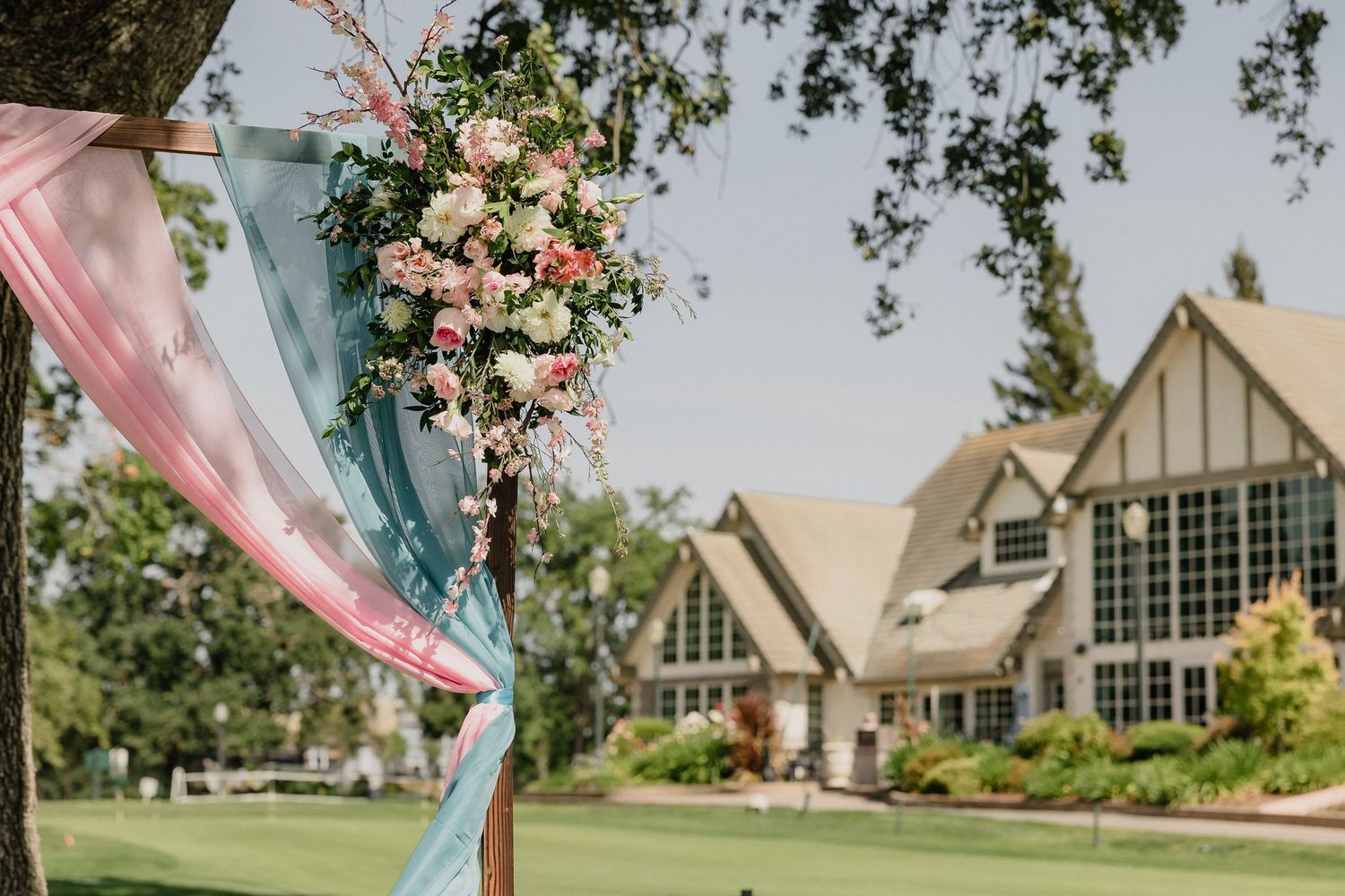 Elegant floral arrangement with pink and white roses decorates outdoor wedding ceremony with tudor style house in background.