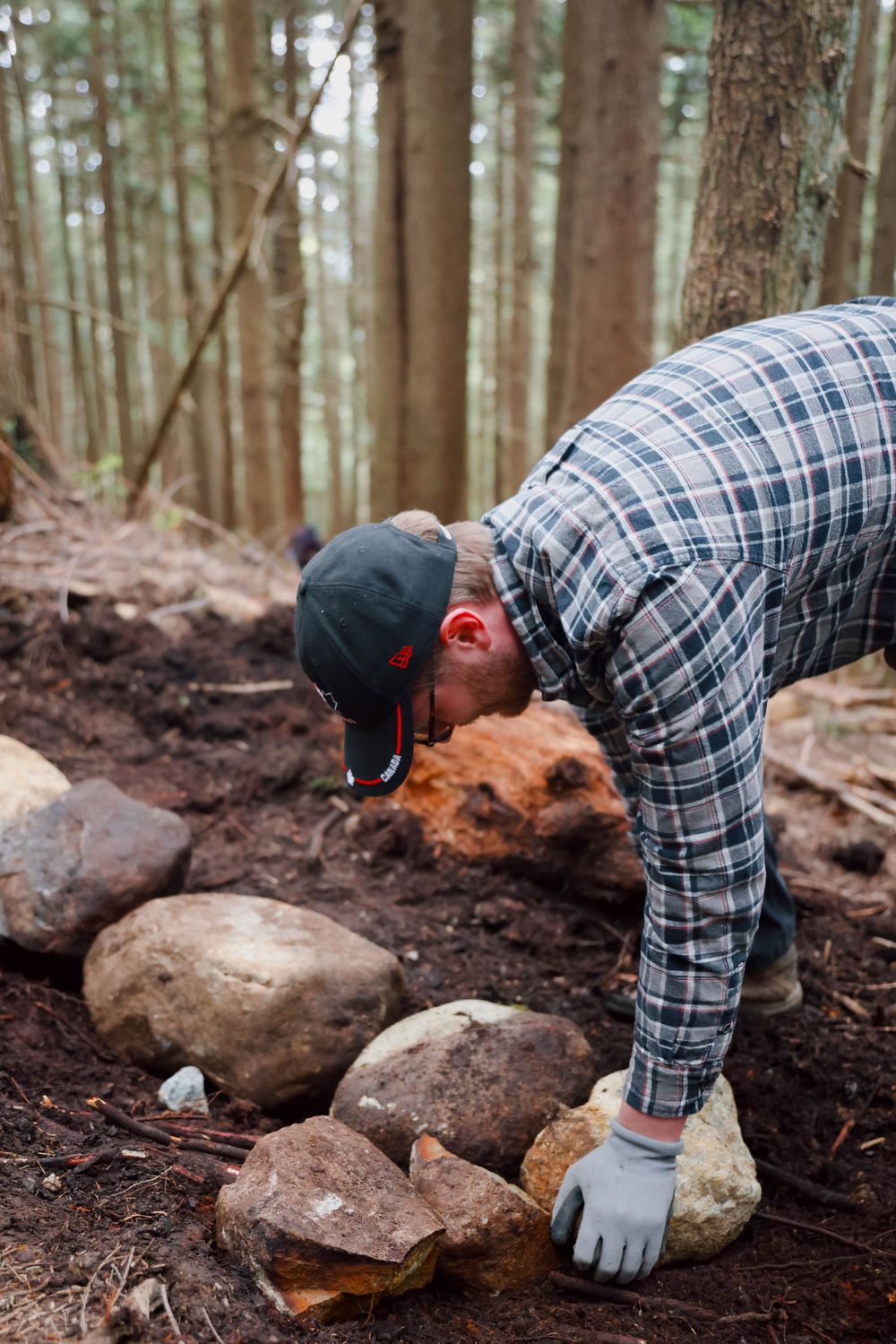 Someone in plaid shirt and cap bends down to pick up rocks while trail building in a dense forest.