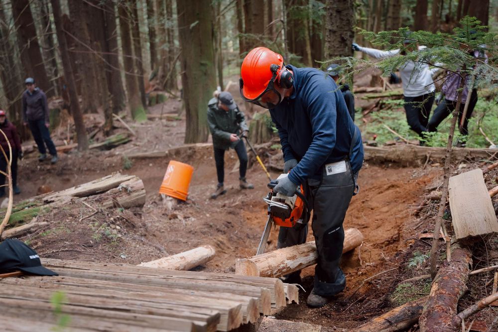 Trail builder wearing safety gear operates chainsaw on wooden features while maintaining a mountain bike trail in forest.