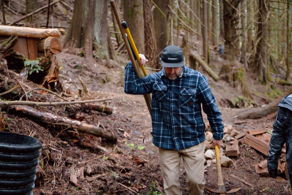 A hiker wearing a blue plaid shirt and headlamp carries tools while walking through a forested trail.