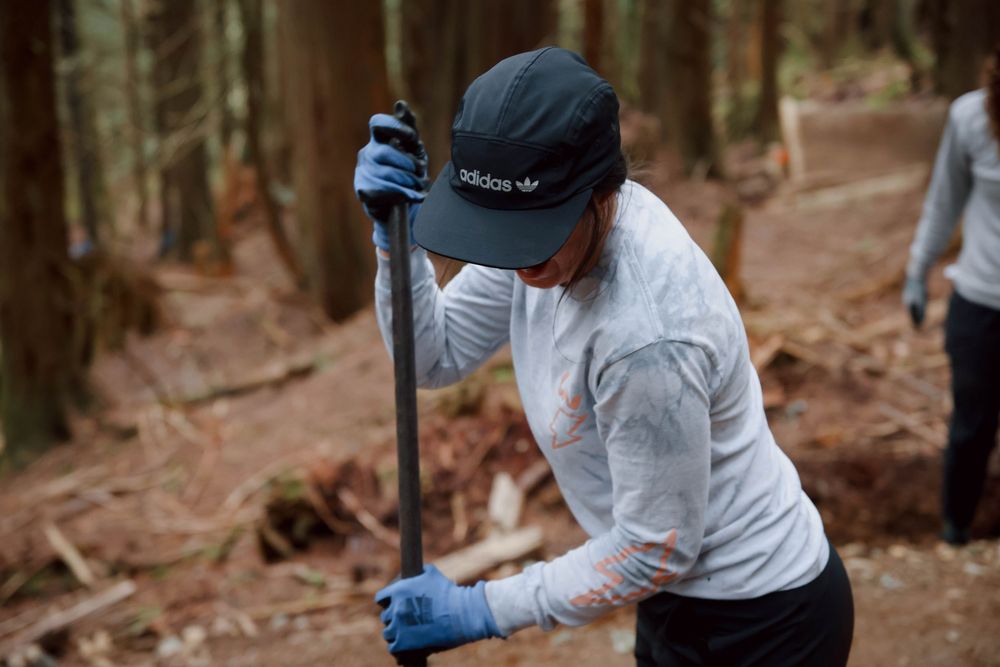 Person wearing blue gloves and white jacket cleans trail with tool in forest setting during volunteer work.