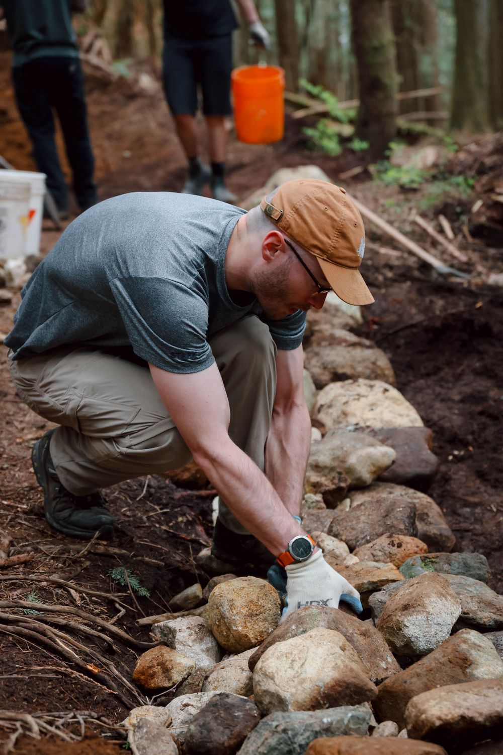 Trail worker in khaki pants and baseball cap arranging rocks to build a sustainable hiking trail in forest setting.