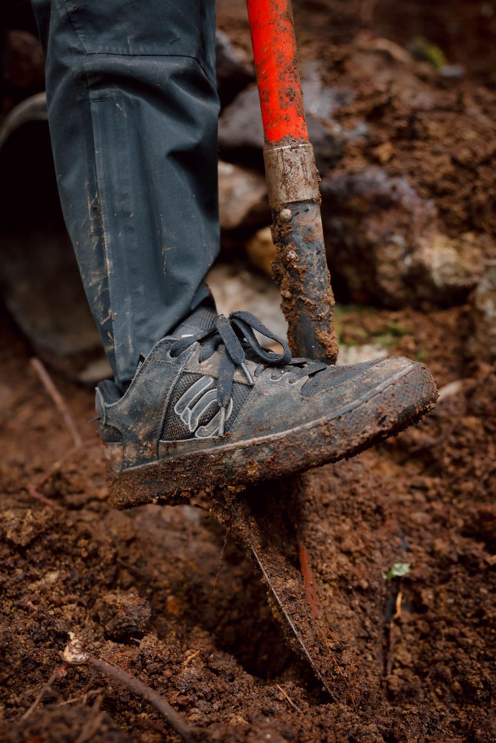 A muddy work boot steps on a shovel while digging in dark soil during garden or construction work.