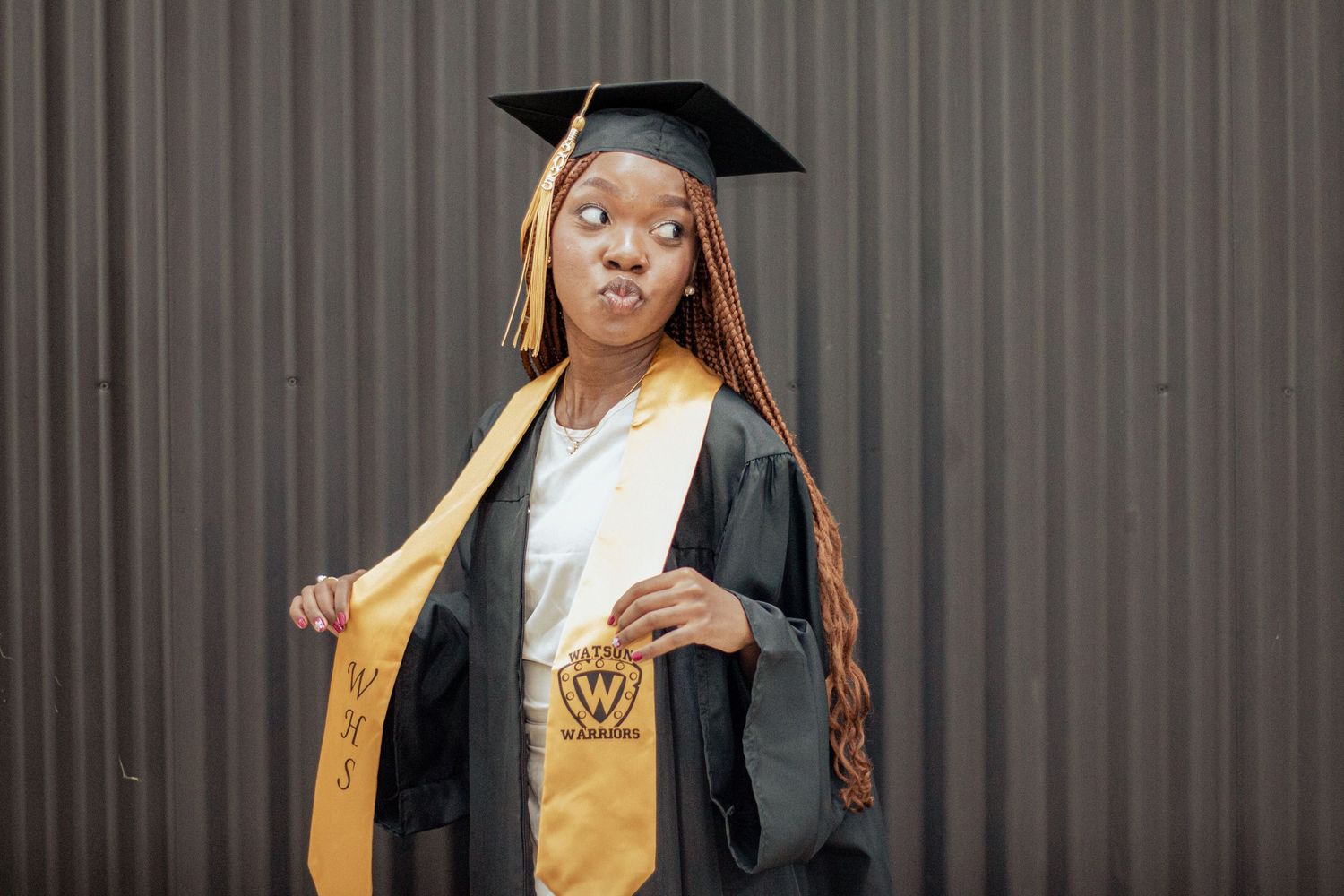 Graduate in black and gold academic regalia poses playfully against gray wall background.