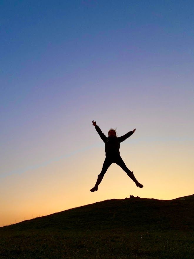 Silhouette of person jumping joyfully against colorful sunset sky on hilltop.