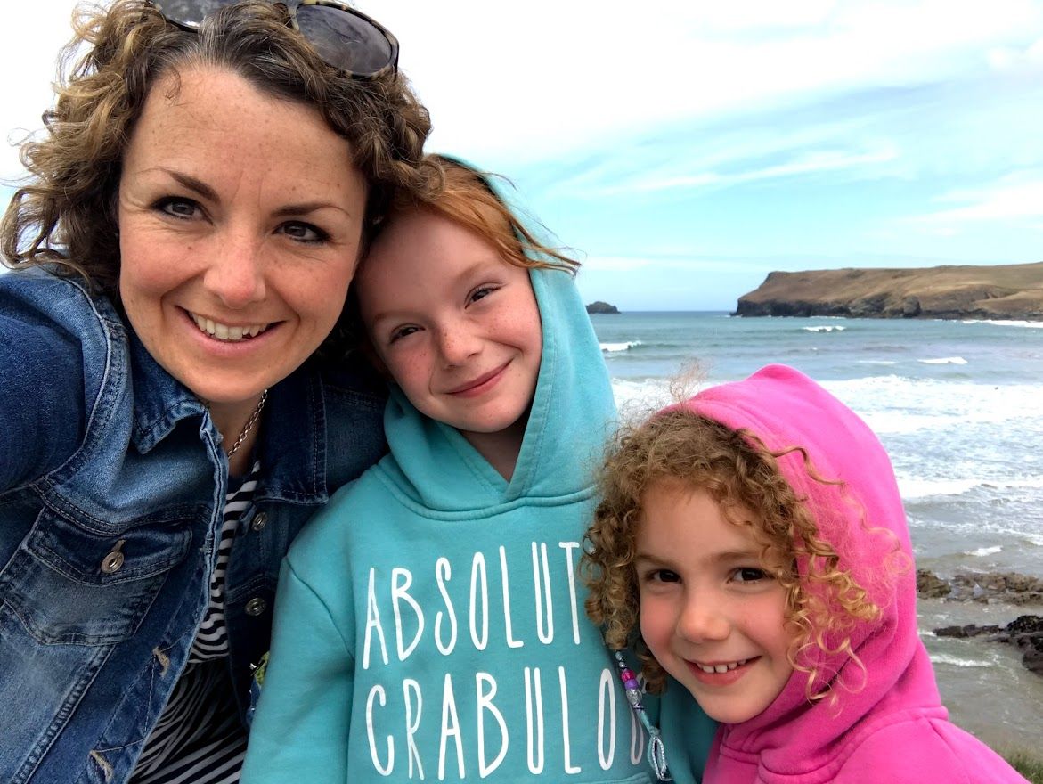Three people in hooded jackets pose for a selfie at a rocky beach with ocean waves and cliffs in the background.