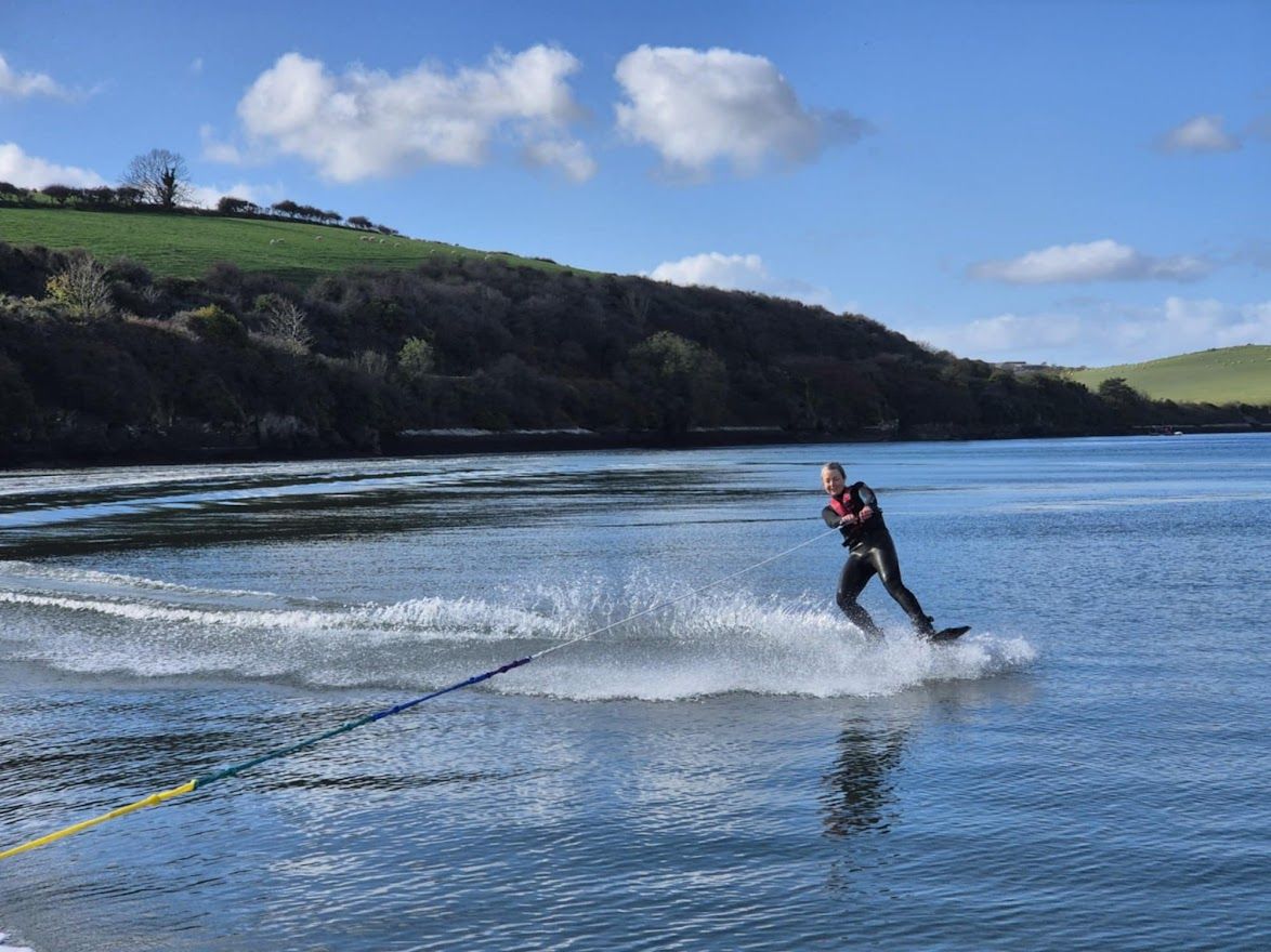 Someone wakeboarding on a blue lake surrounded by green rolling hills on a sunny day with scattered clouds.